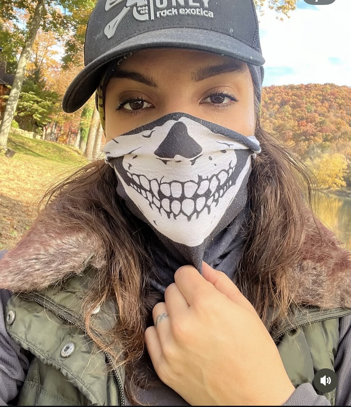 Person wearing a cap and a face mask with a skull design outdoors, with trees and autumn foliage in background.