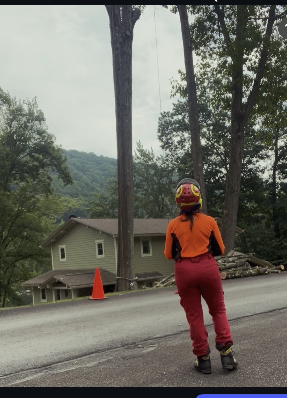 Person wearing an orange jacket, red pants, and a helmet standing on the side of a road with trees and a house in the background.