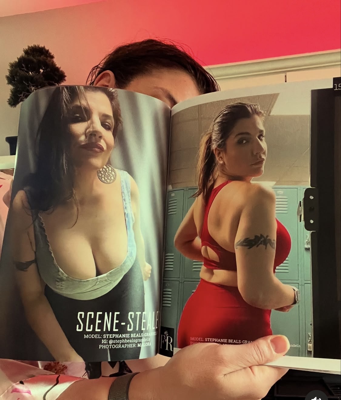 Person holding open magazine with two women in revealing outfits, one in a light top and the other in a red dress, in front of lockers.