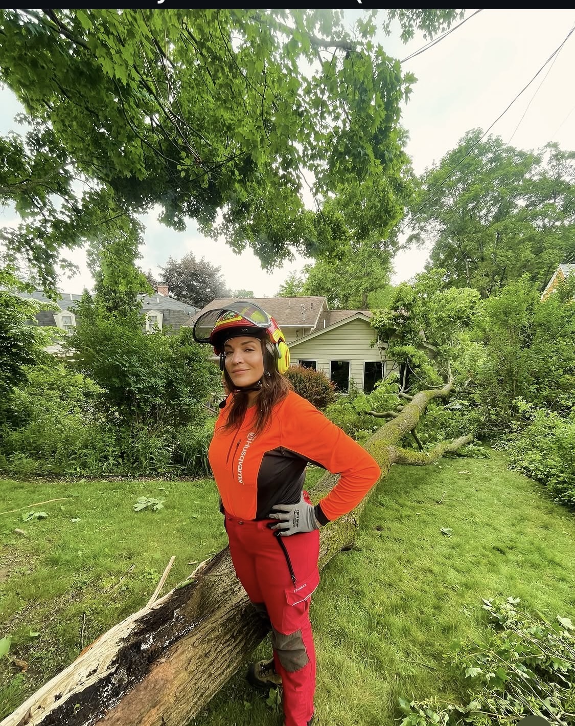 Woman in orange jacket and red pants standing outdoors next to fallen tree, wearing a helmet and gloves, with green trees and house in background.