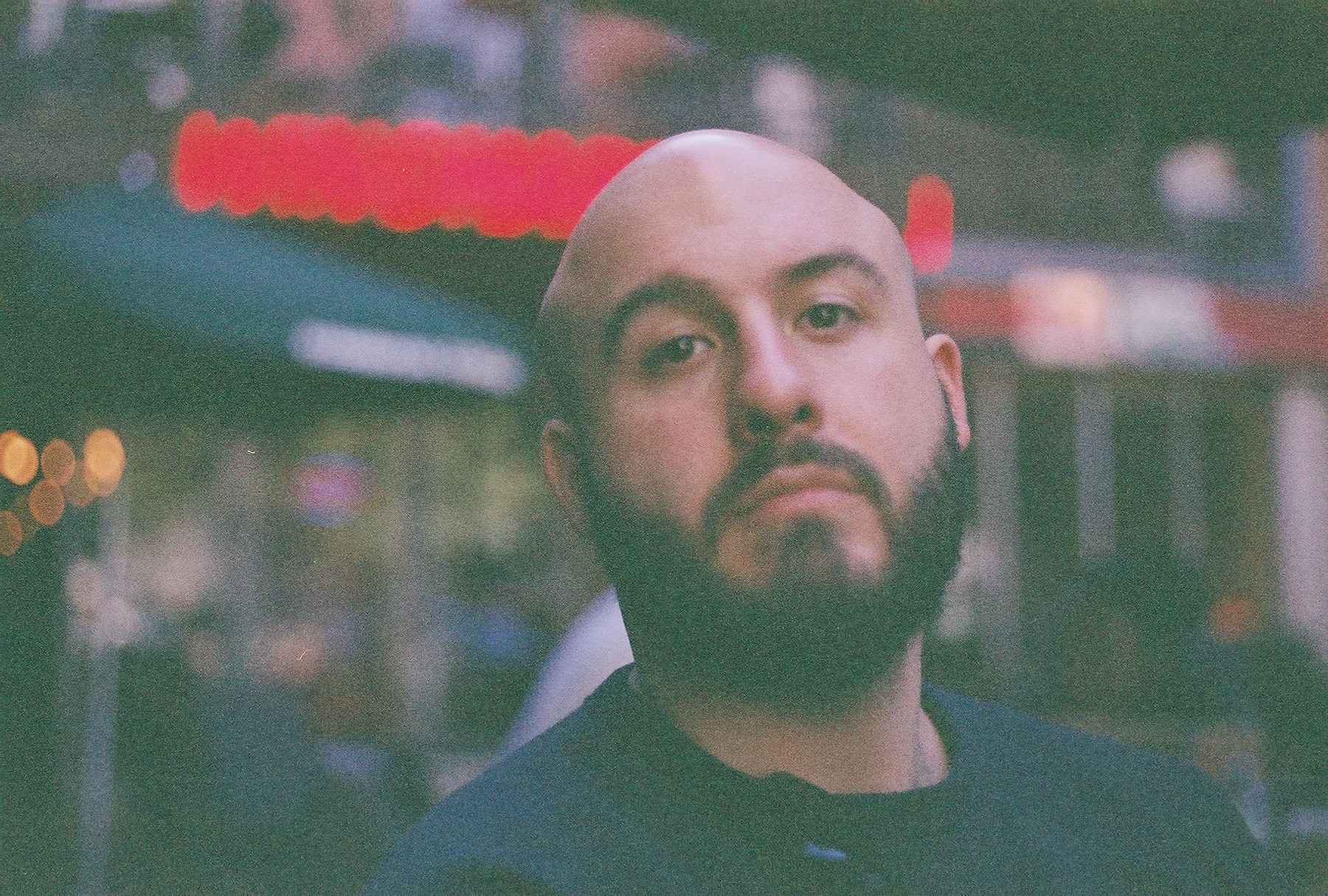 Portrait of a man with a beard and shaved head outdoors, blurred background, neutral expression.