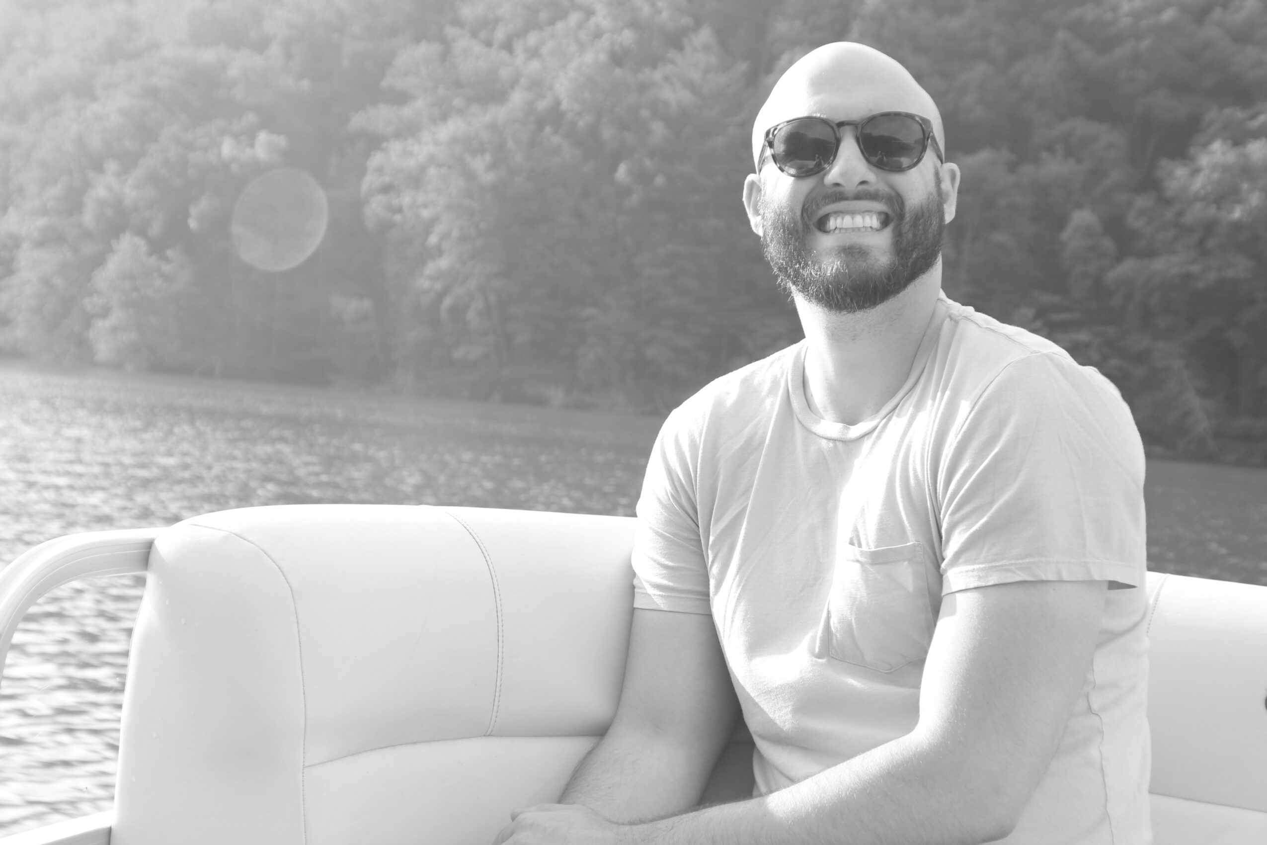 Man with sunglasses smiling on boat near water, trees in background