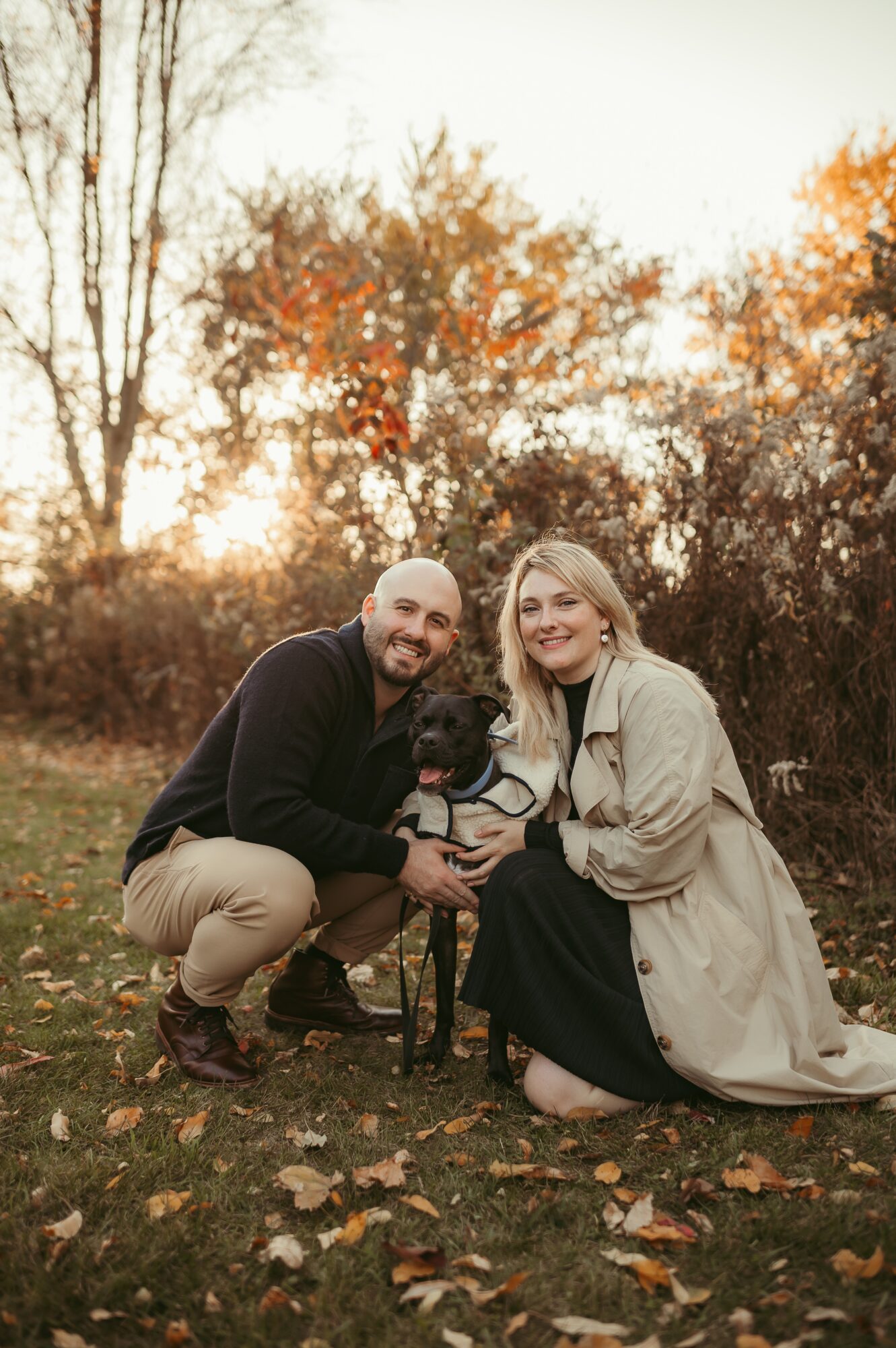 Two people and a dog outdoors in autumn, with trees and fallen leaves, smiling at the camera.