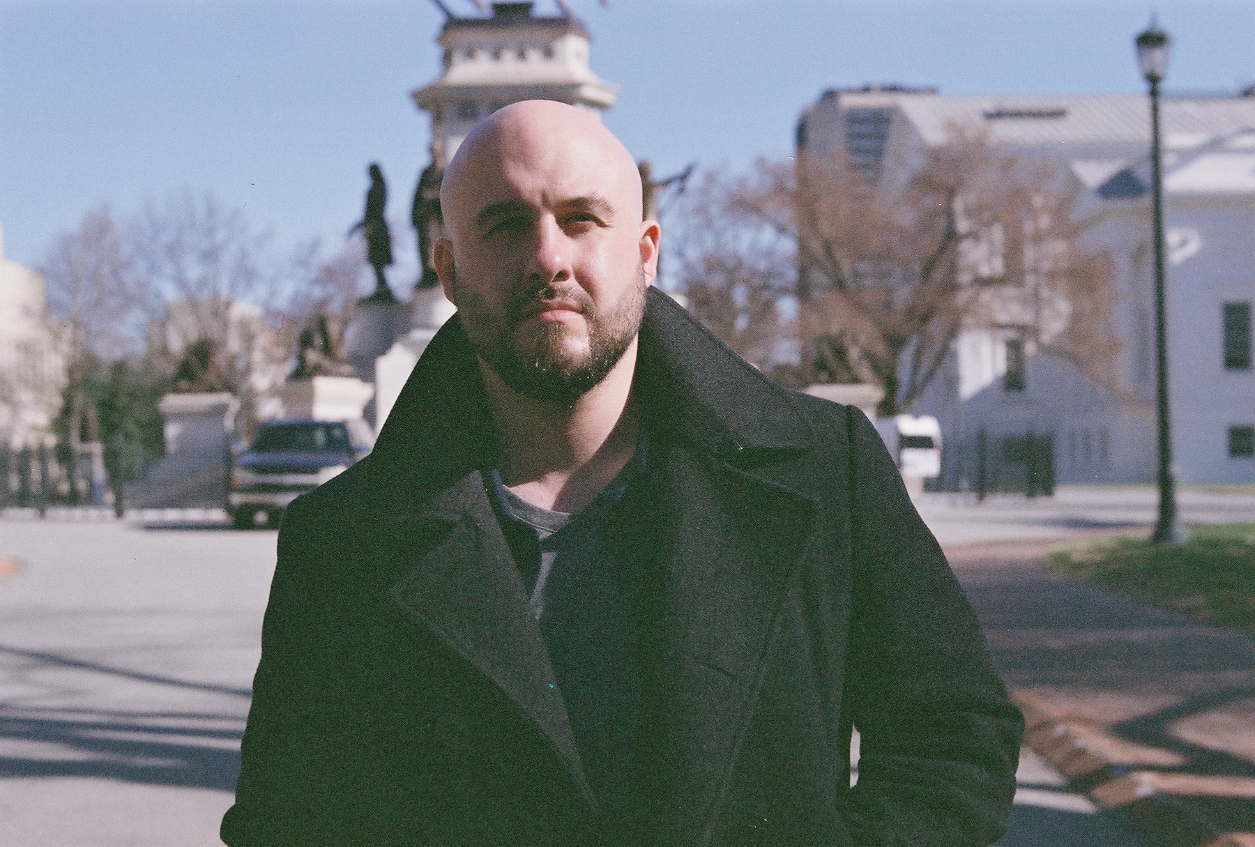 Man with a beard and bald head wearing a black coat outdoors on a sunny day.