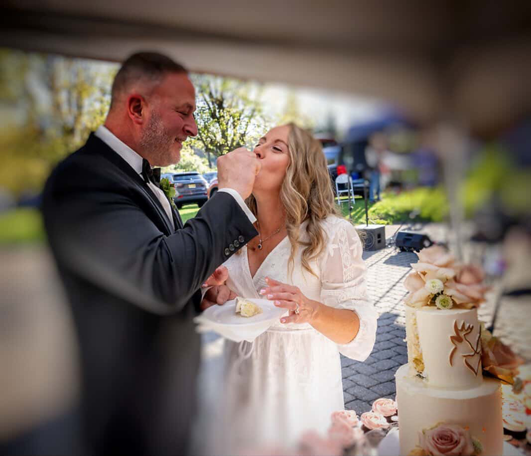 A couple at a wedding cake cutting, smiling, outdoors with trees and cars in background.