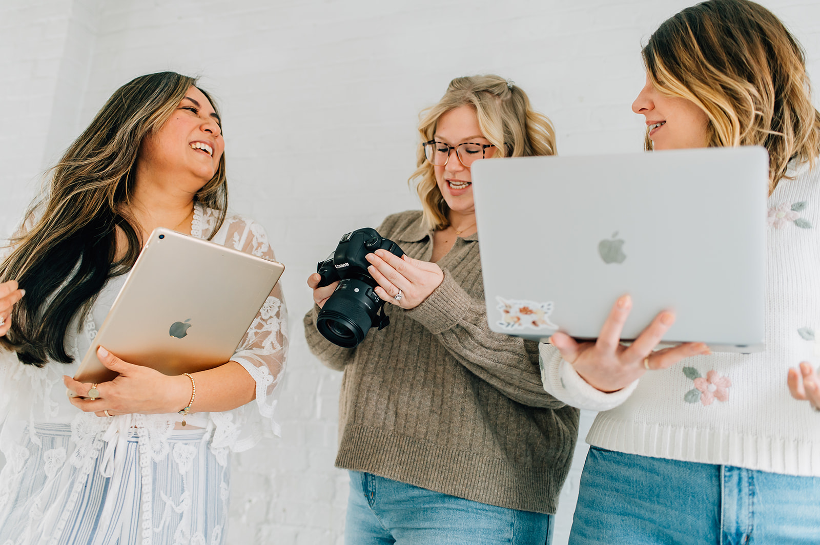 Three women smiling and talking, one holding a camera, two holding laptops, against a plain background.