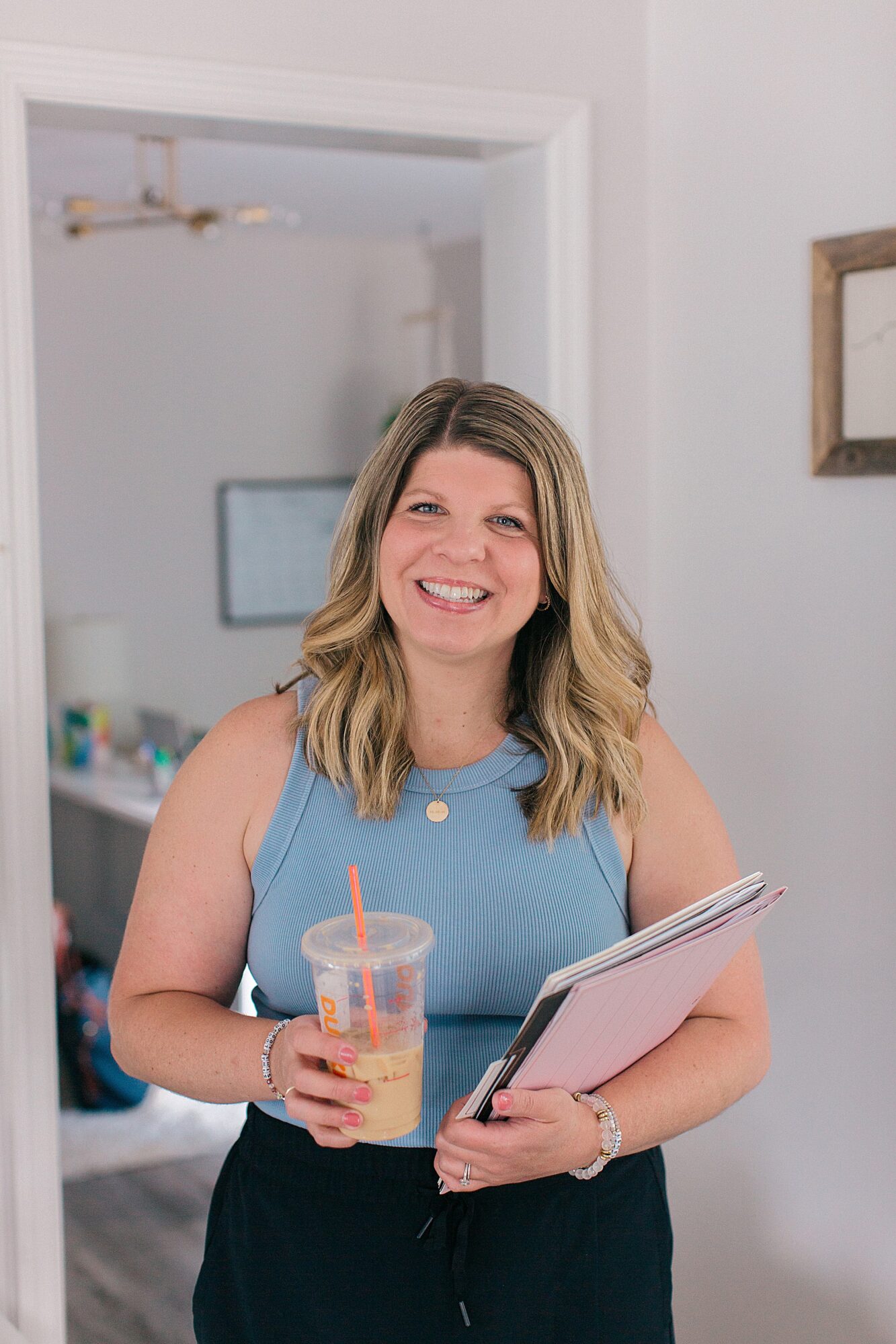 Smiling woman with shoulder-length hair holding a drink and a notebook in a bright room.