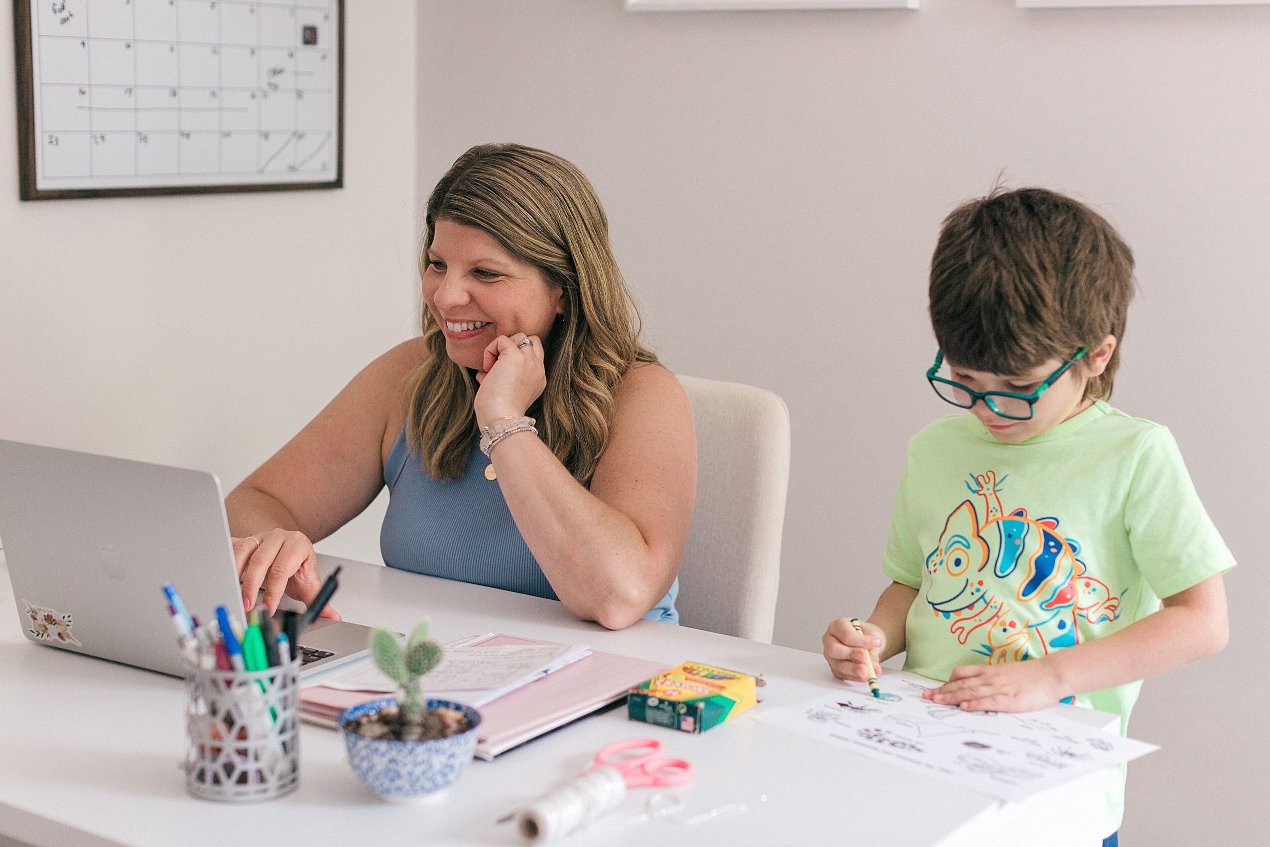 Woman and child sitting at a desk with a laptop, papers, and stationery, smiling and working together in a bright room.