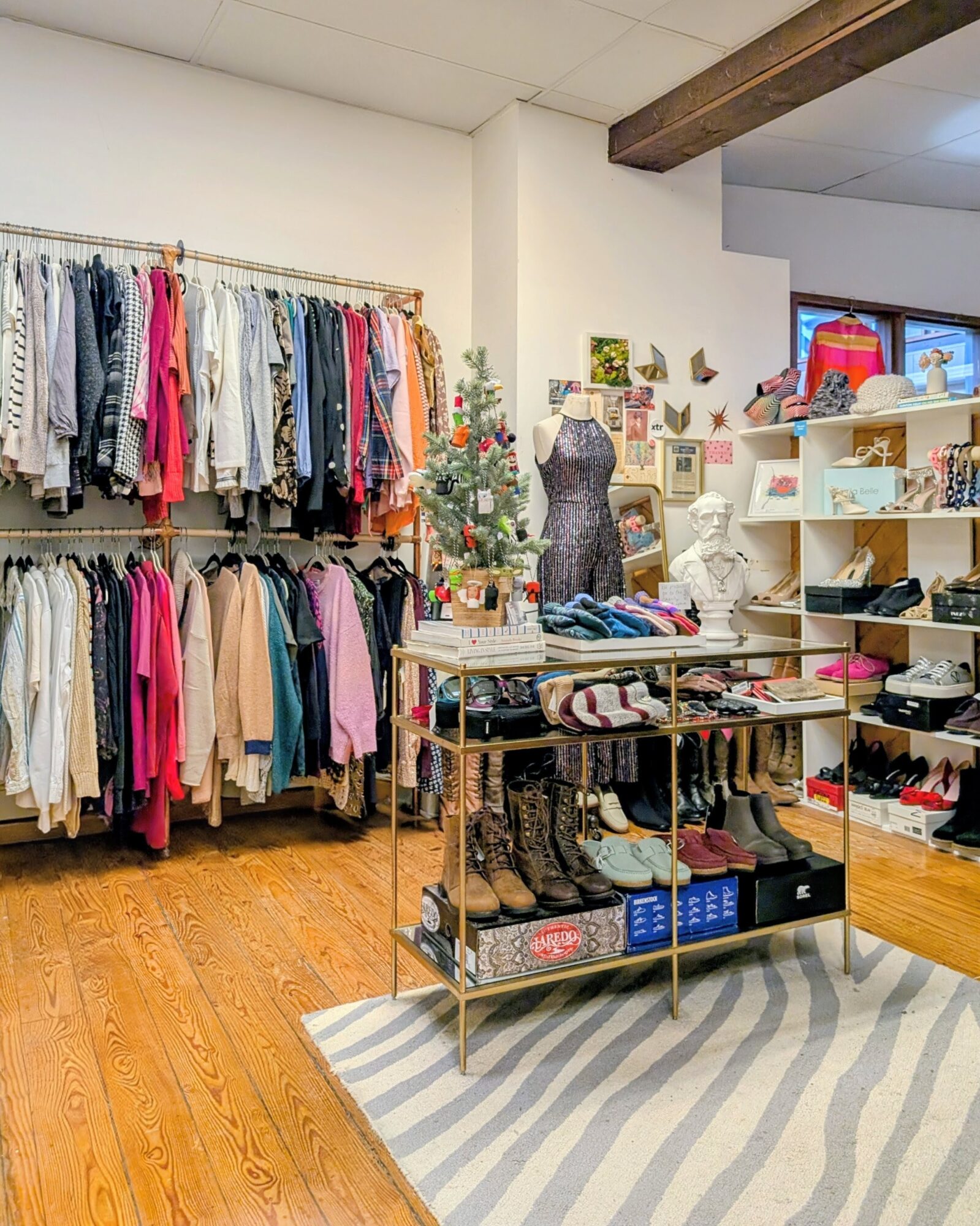 Clothing store with racks of colorful clothes, shelves with accessories, and a mannequin, wooden floor, and striped rug.