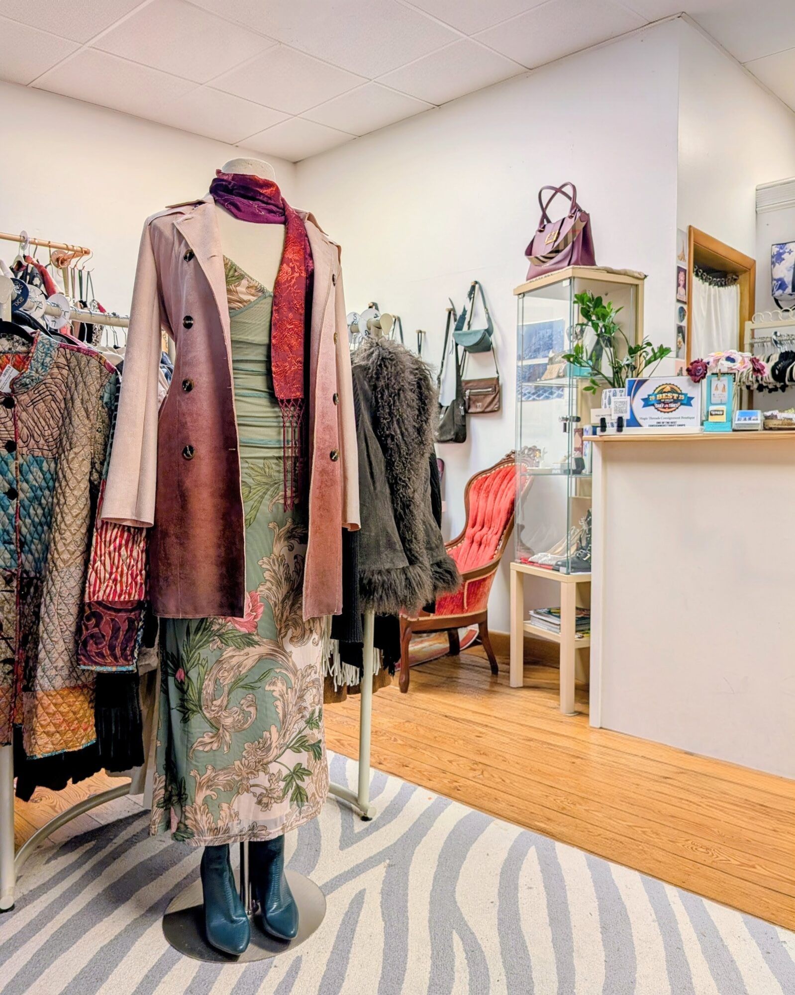 Clothing display with a mannequin wearing a long coat, scarf, and skirt inside a boutique. Shelves and chairs are visible in the background.