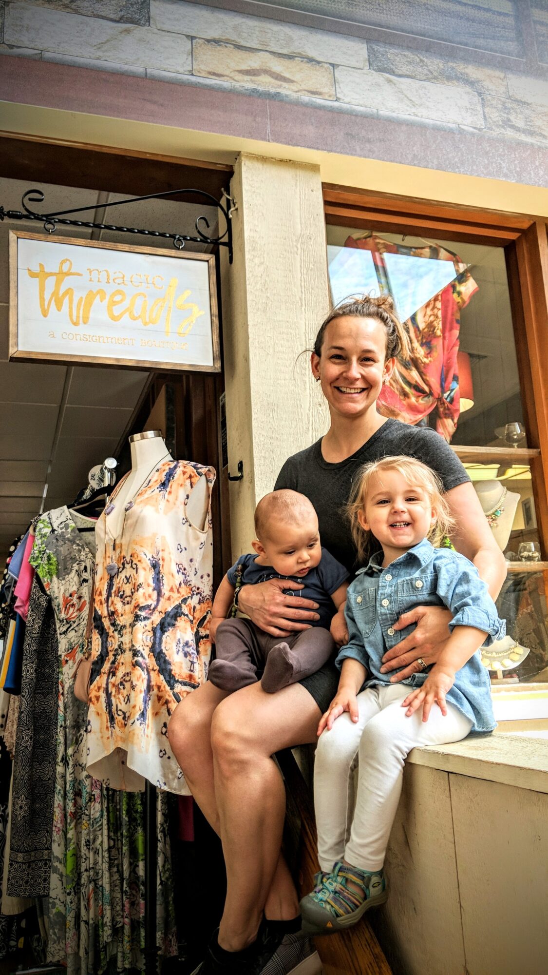 Smiling woman holding two children outside a shop, one child sitting on her lap and the other standing beside her.