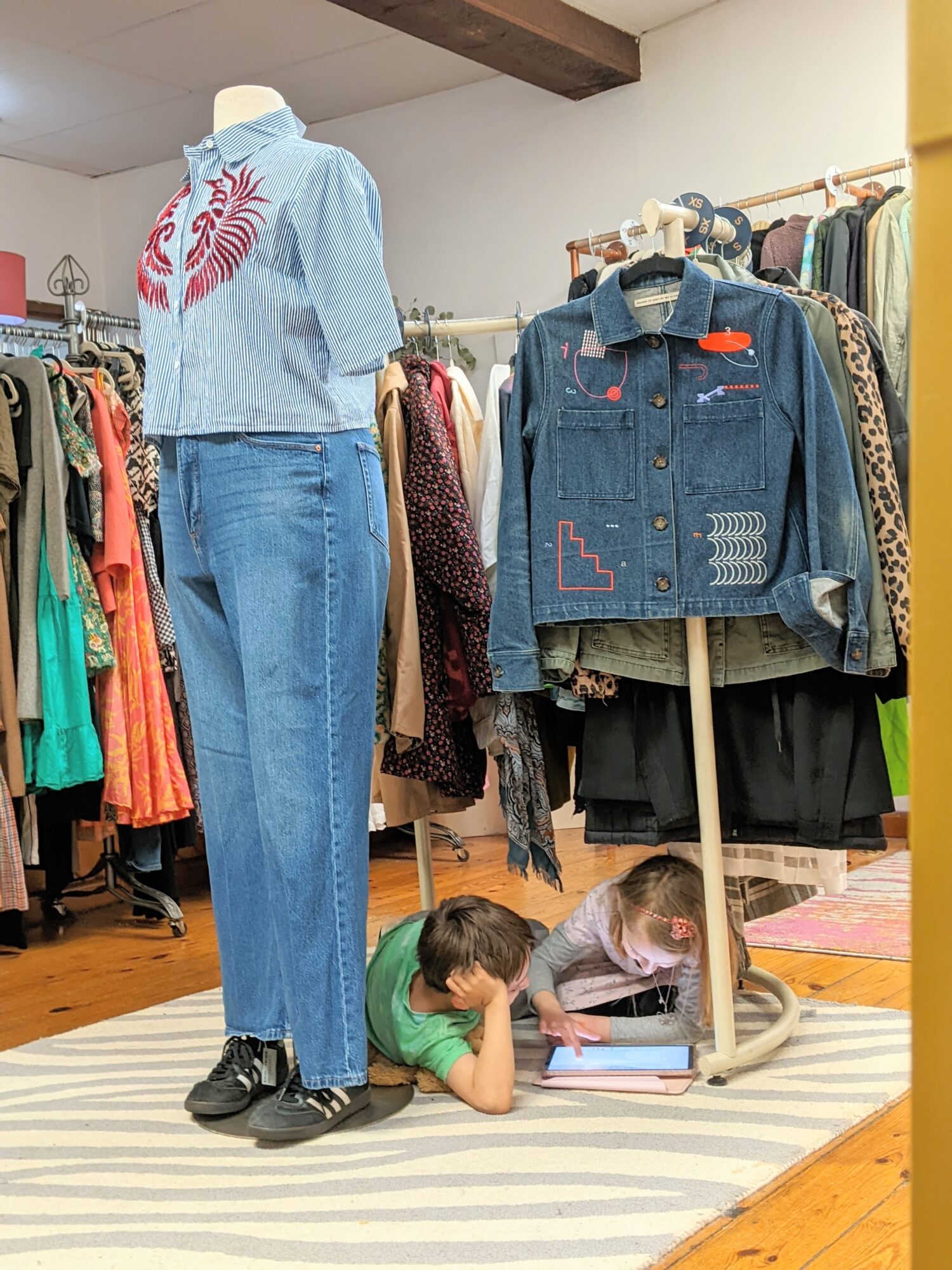 Two children lie on the floor reading books beneath a mannequin dressed in a blue shirt and jeans, with clothing racks nearby.
