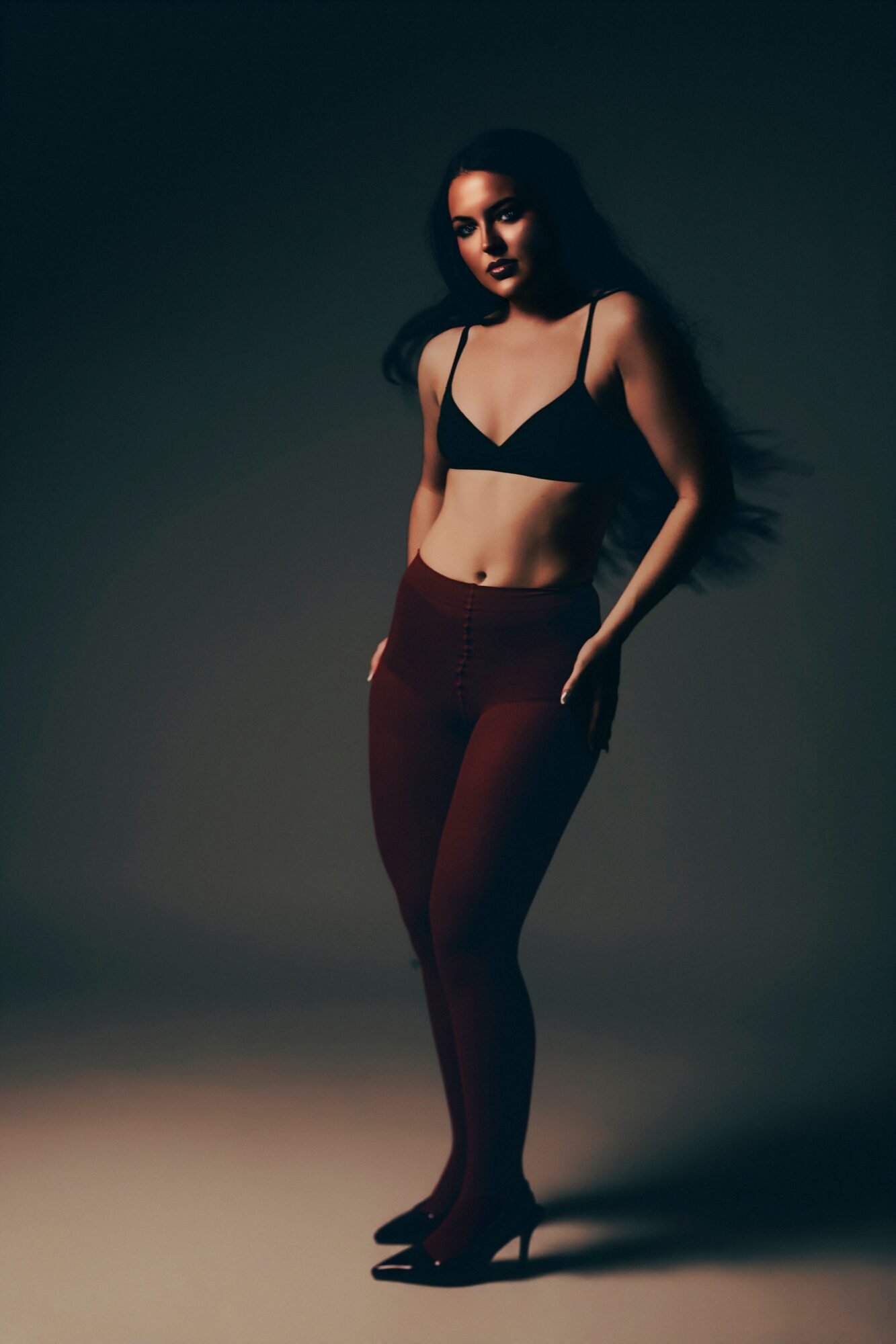 Woman in black top and red pants standing with hand in pocket, dark background, dramatic lighting.