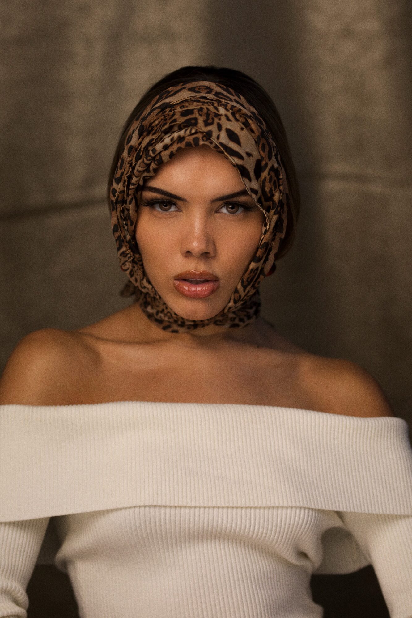 Woman with a leopard print headscarf, off-shoulder white top, neutral background, serious expression.