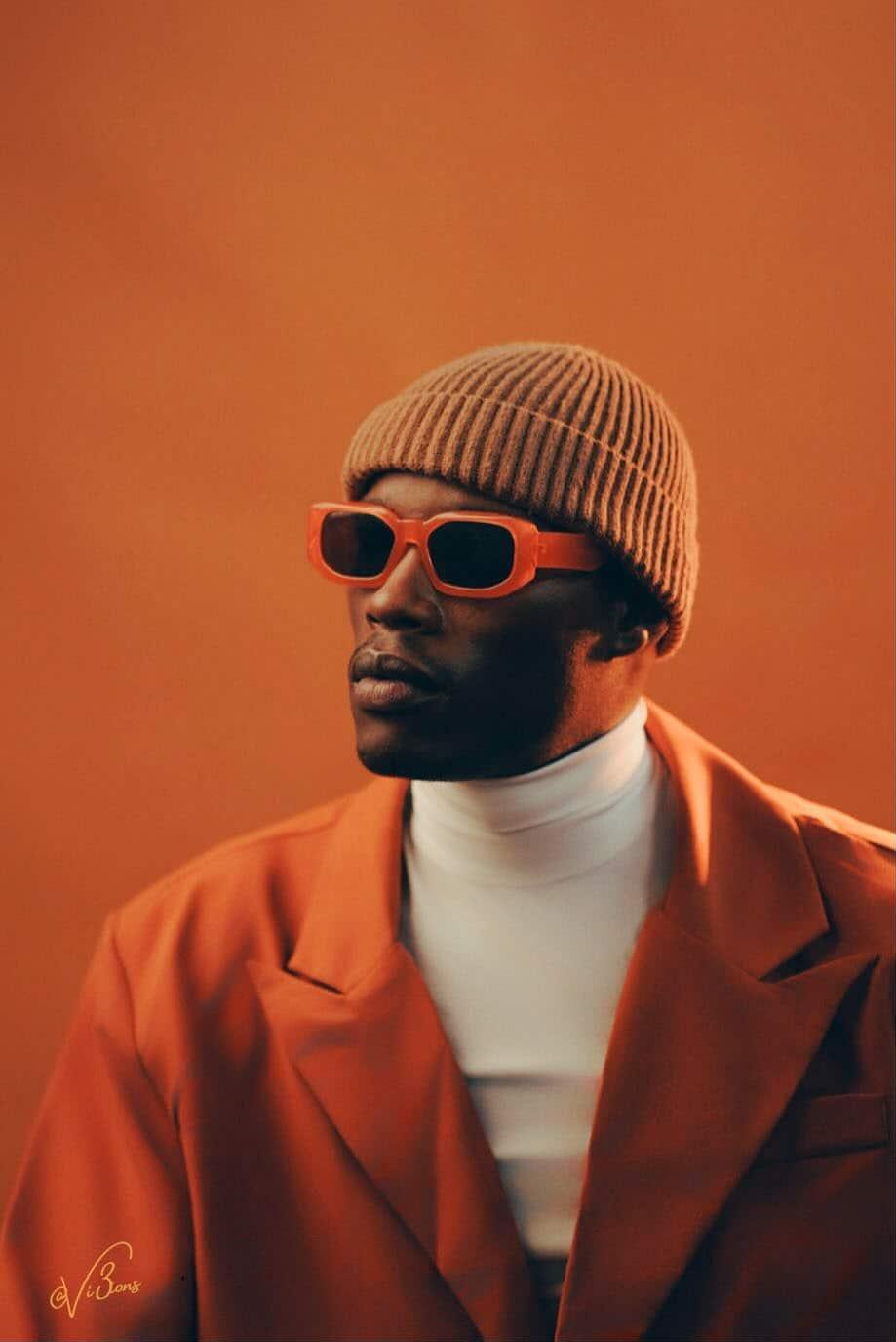 Portrait of a person wearing a beige knit hat, orange sunglasses, an orange blazer, and a white turtleneck against an orange background.