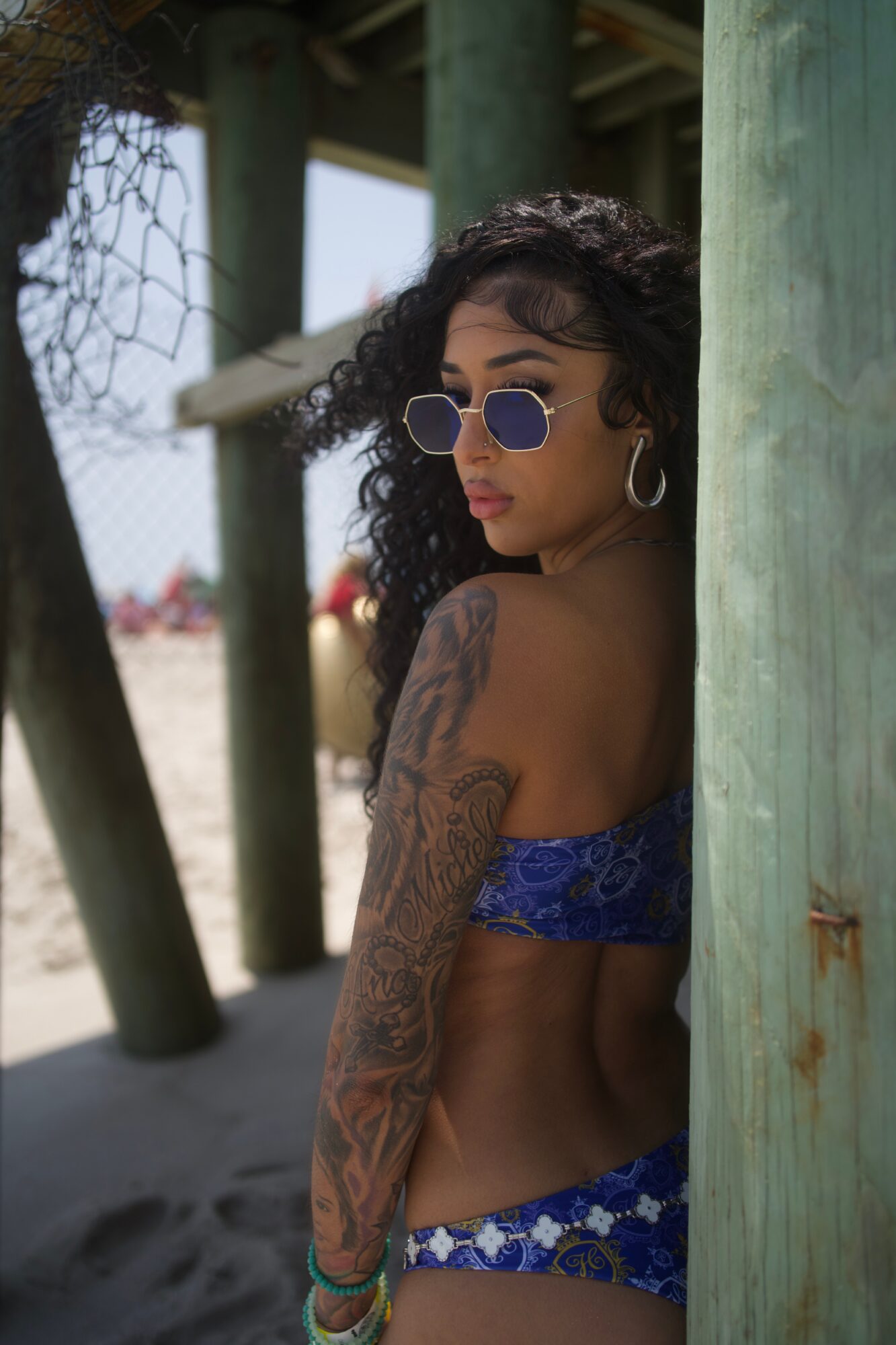 Woman with curly hair wearing sunglasses and a blue patterned bikini at the beach, standing near wooden posts.