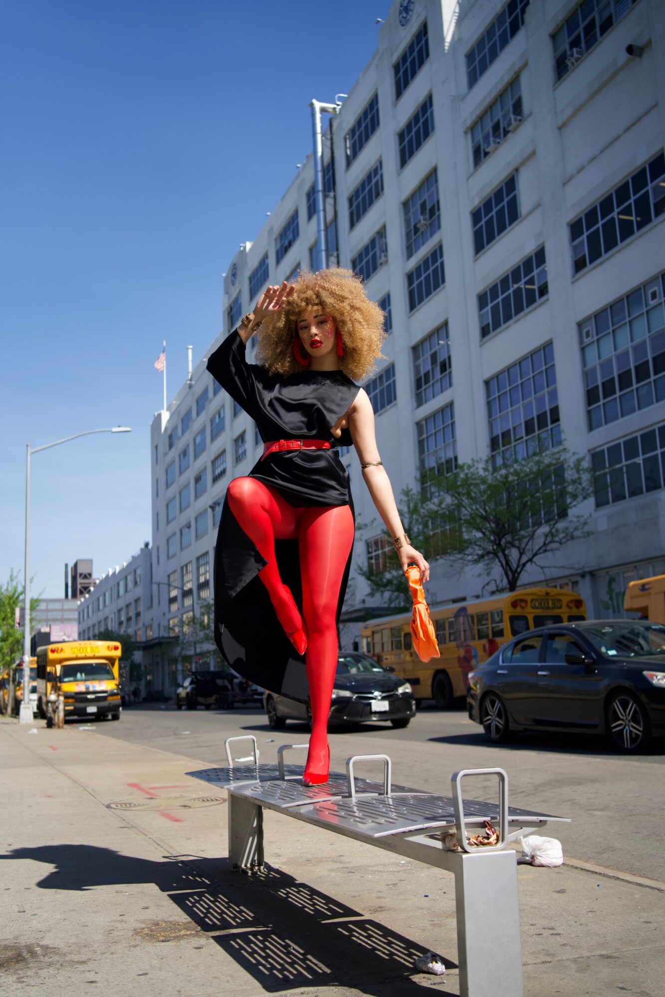 Person with curly hair and red tights standing on a city sidewalk, with tall white buildings and cars in the background.