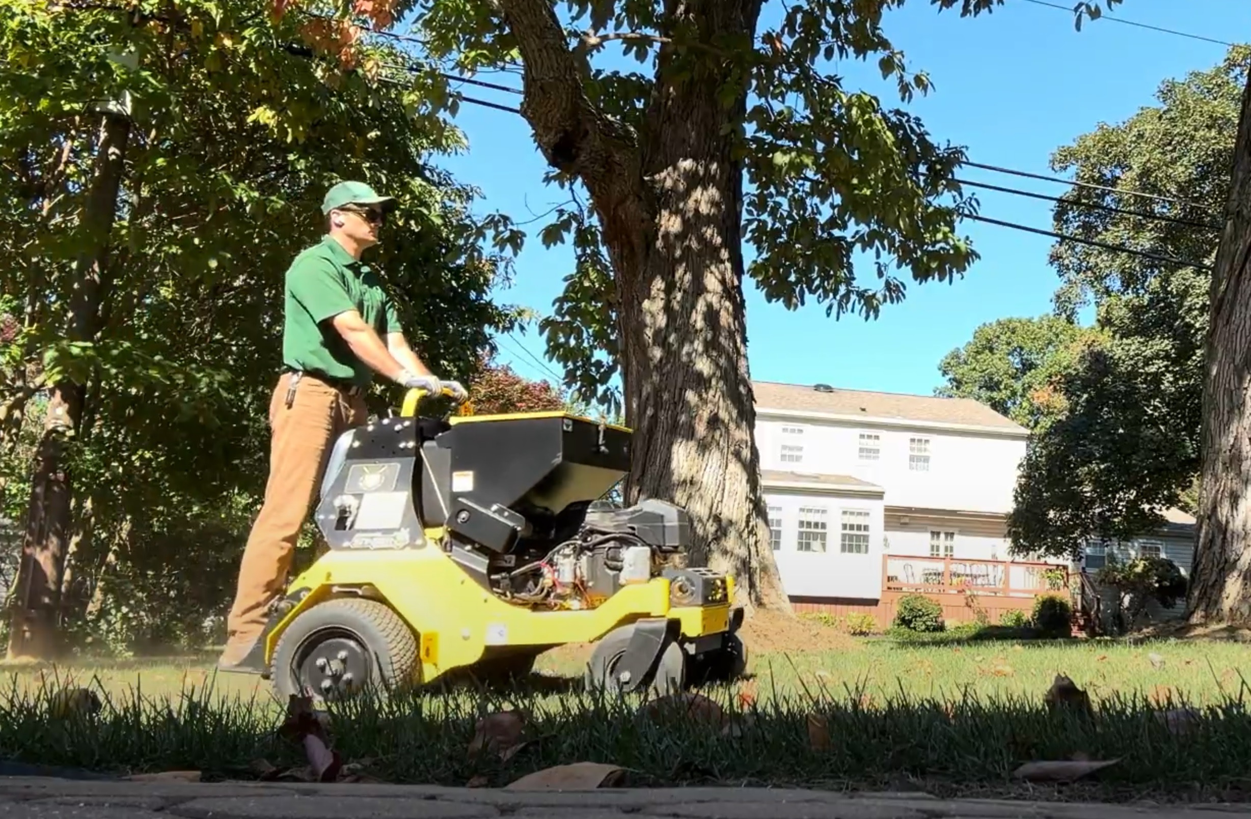 Person operating a yellow and black lawn mower under a large tree outdoors, with buildings and cars in the background.