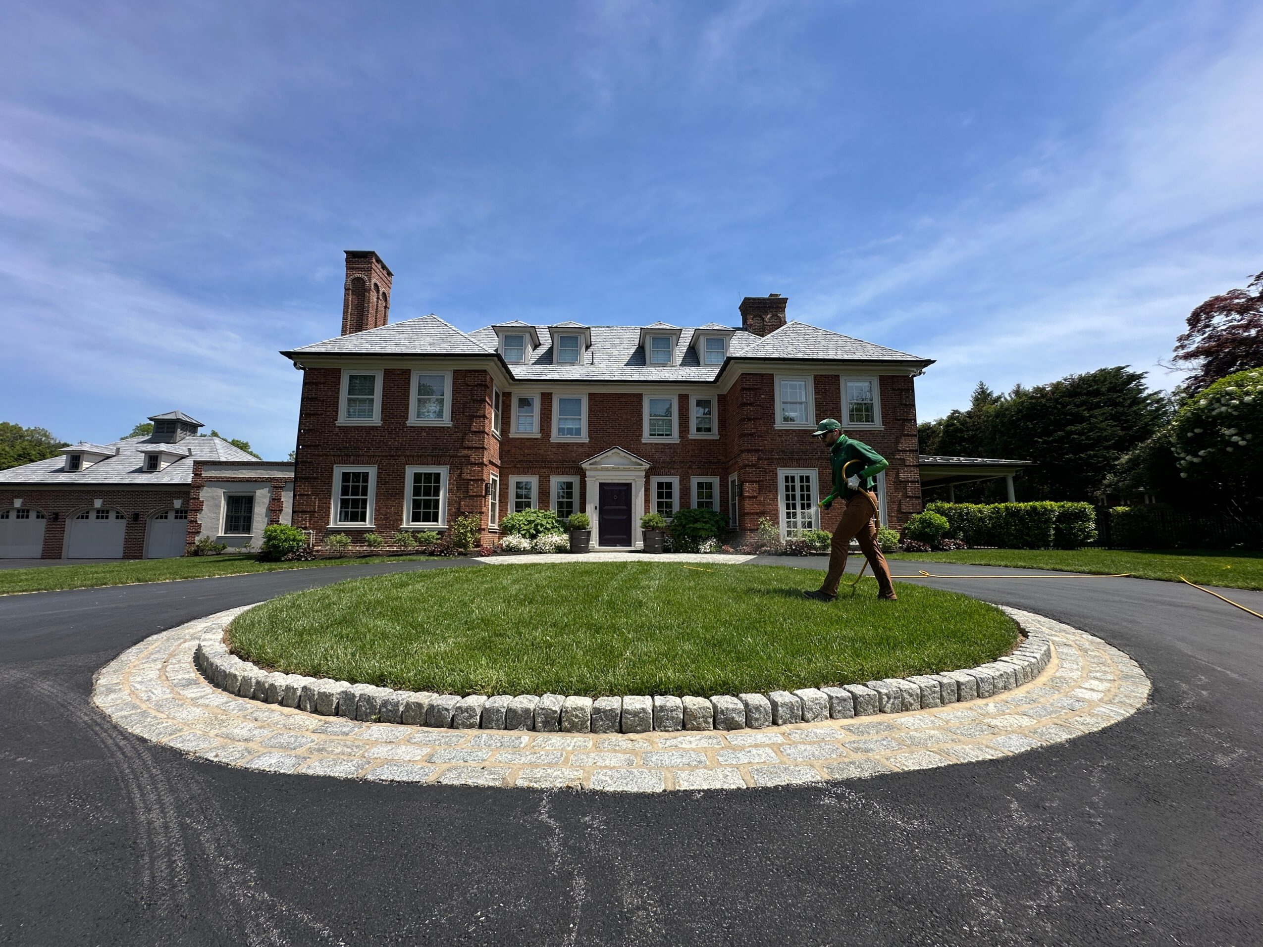 Large brick mansion with a circular driveway and a person walking on the grass in front, under a blue sky.