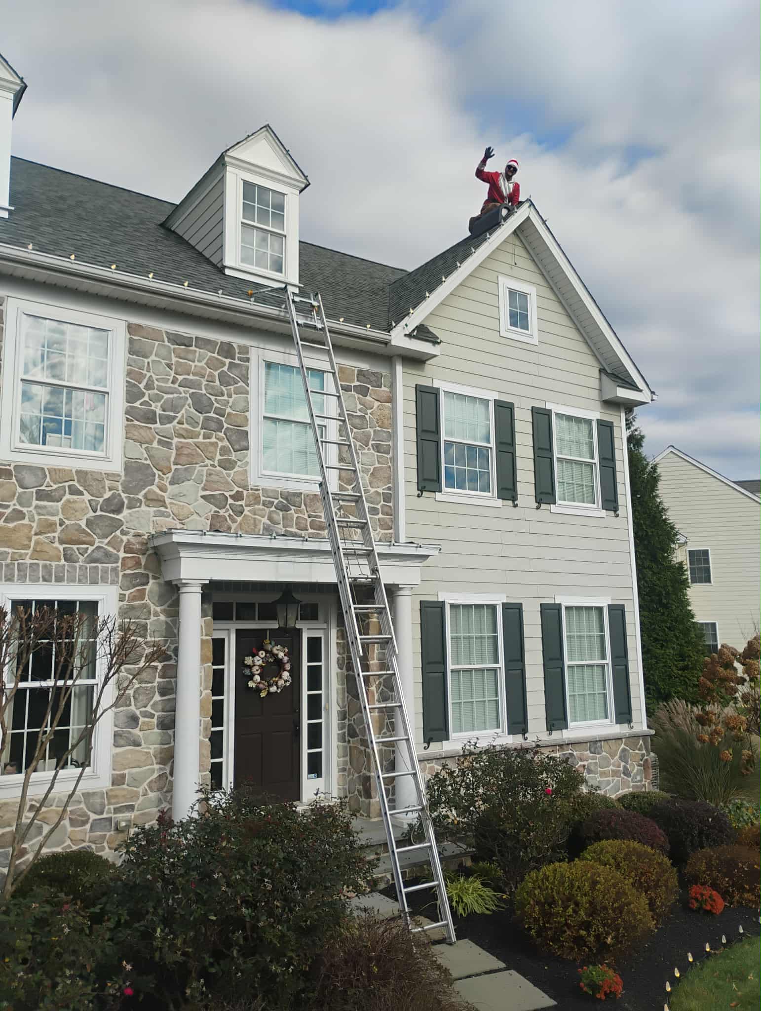 House with stone and siding exterior, ladder leaning against it, Santa Claus figure on roof, cloudy sky, garden in front.