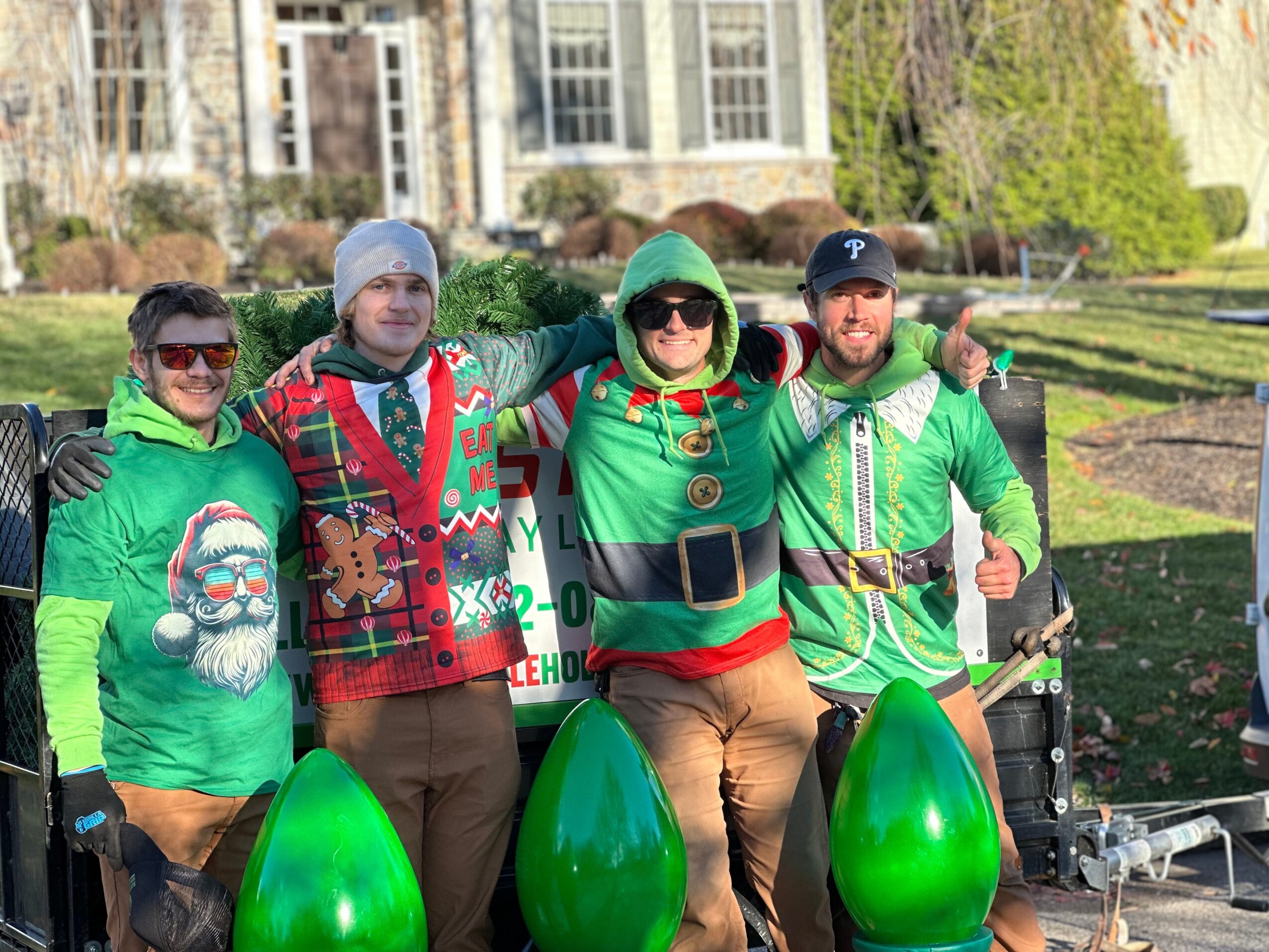 Five people in Christmas-themed costumes stand outdoors, smiling with arms around each other, in front of a sign and trees.
