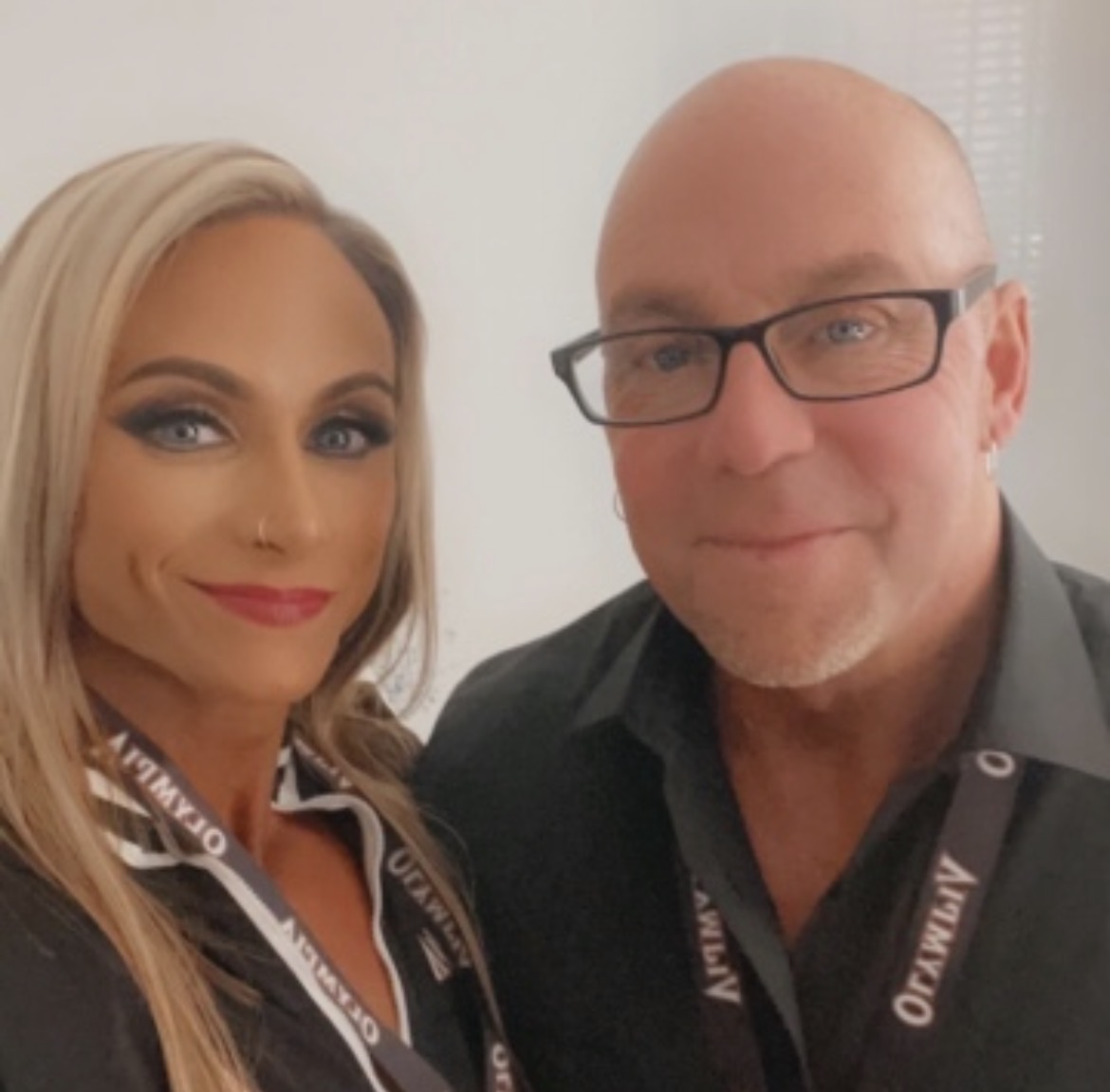 Two people, a woman with blonde hair and makeup, and a man with glasses, pose together indoors.