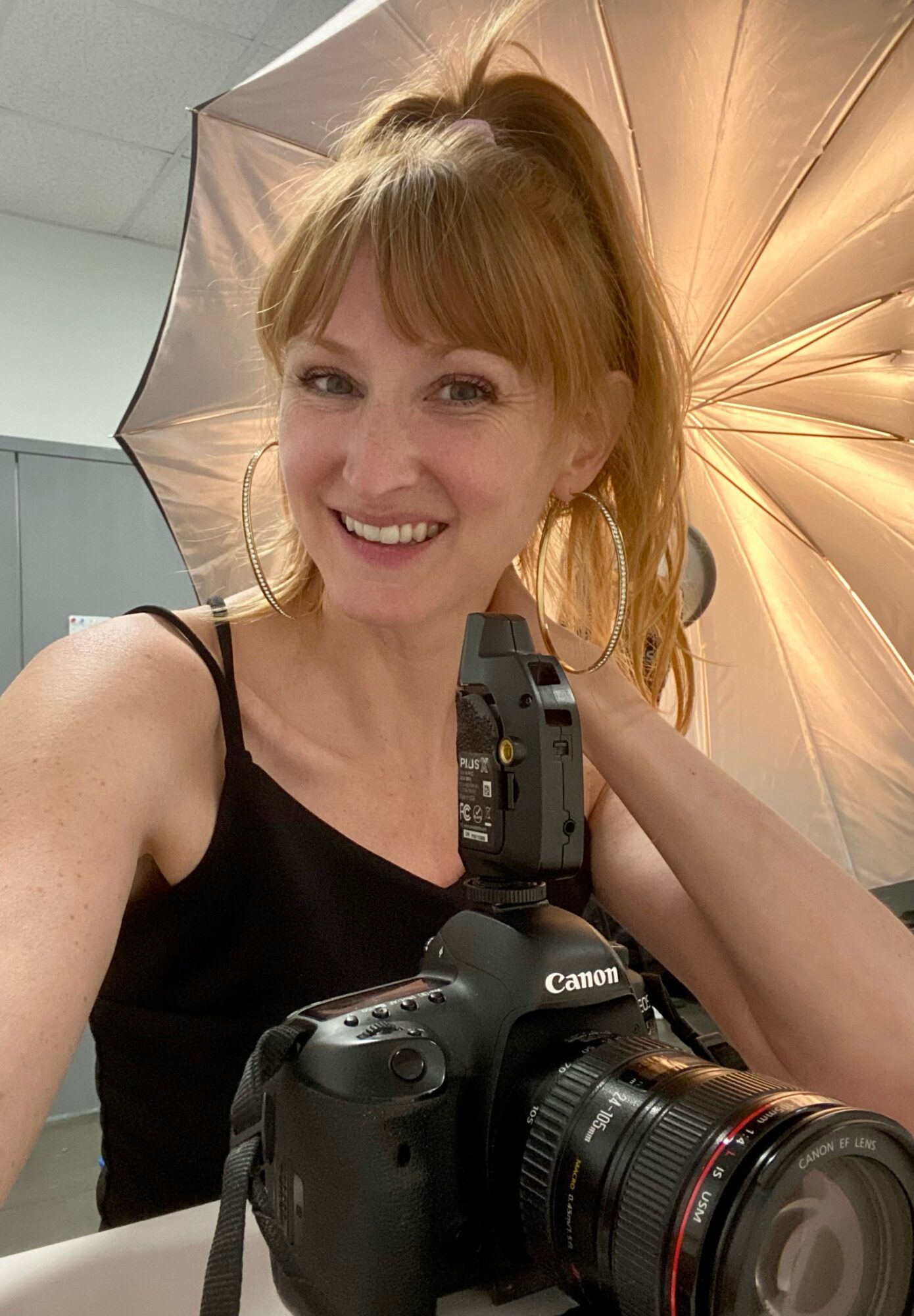 Woman with red hair smiling, holding a camera, in a photography studio with a large umbrella light behind her.