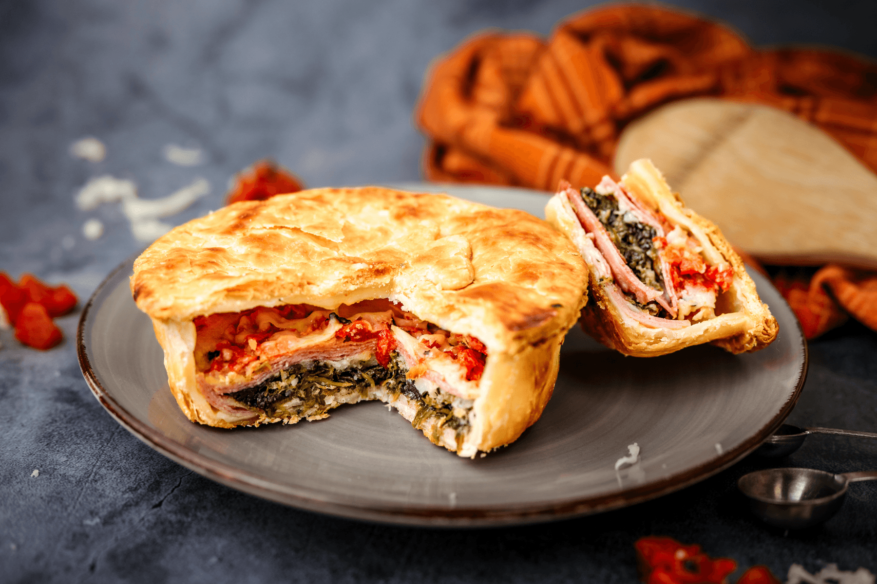 A savory pie with a golden crust, cut open to reveal layers of meat, vegetables, and cheese, on a dark plate.