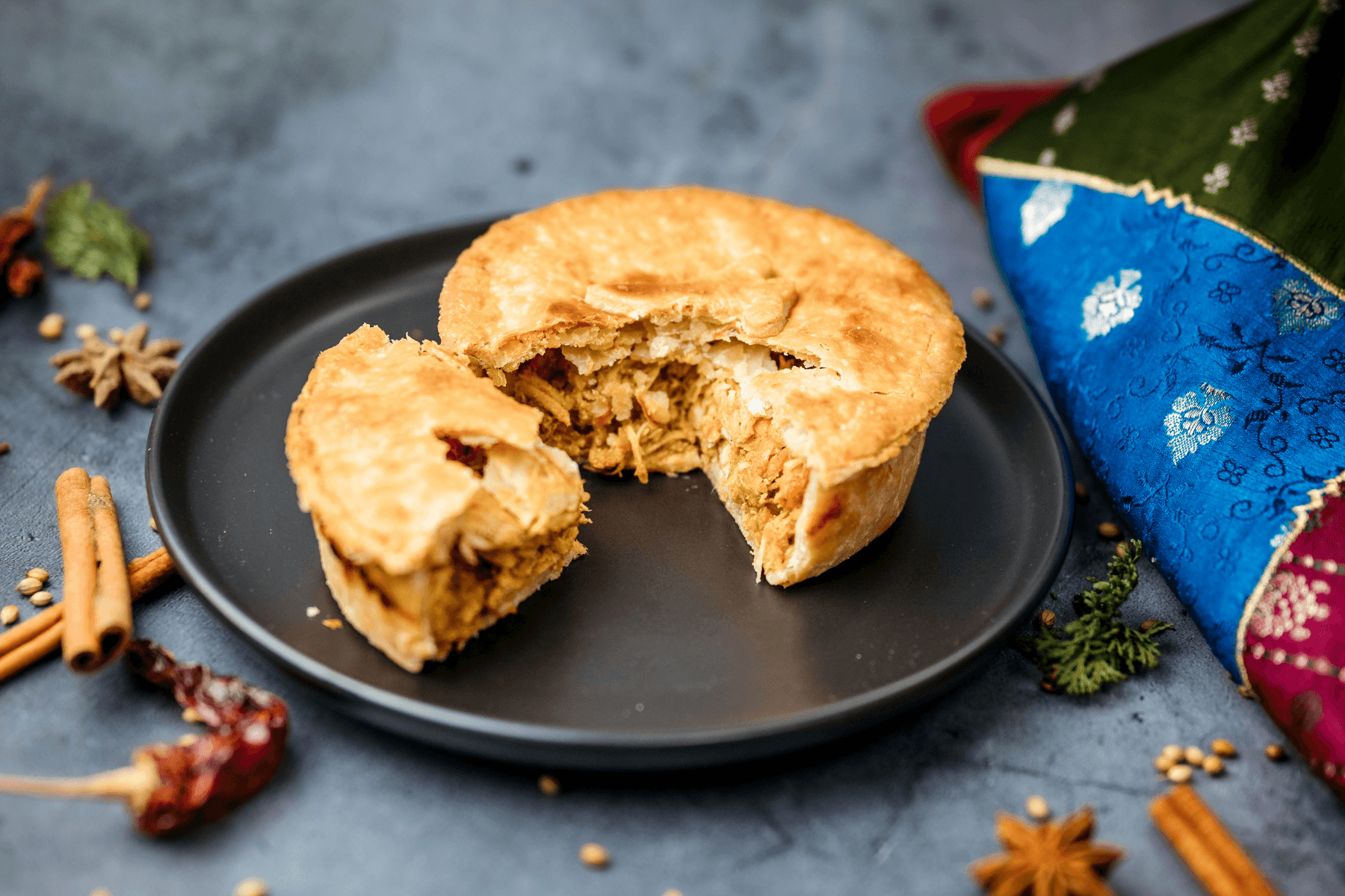 A partially eaten round pastry with a filling on a black plate, with spices and a colorful cloth nearby.
