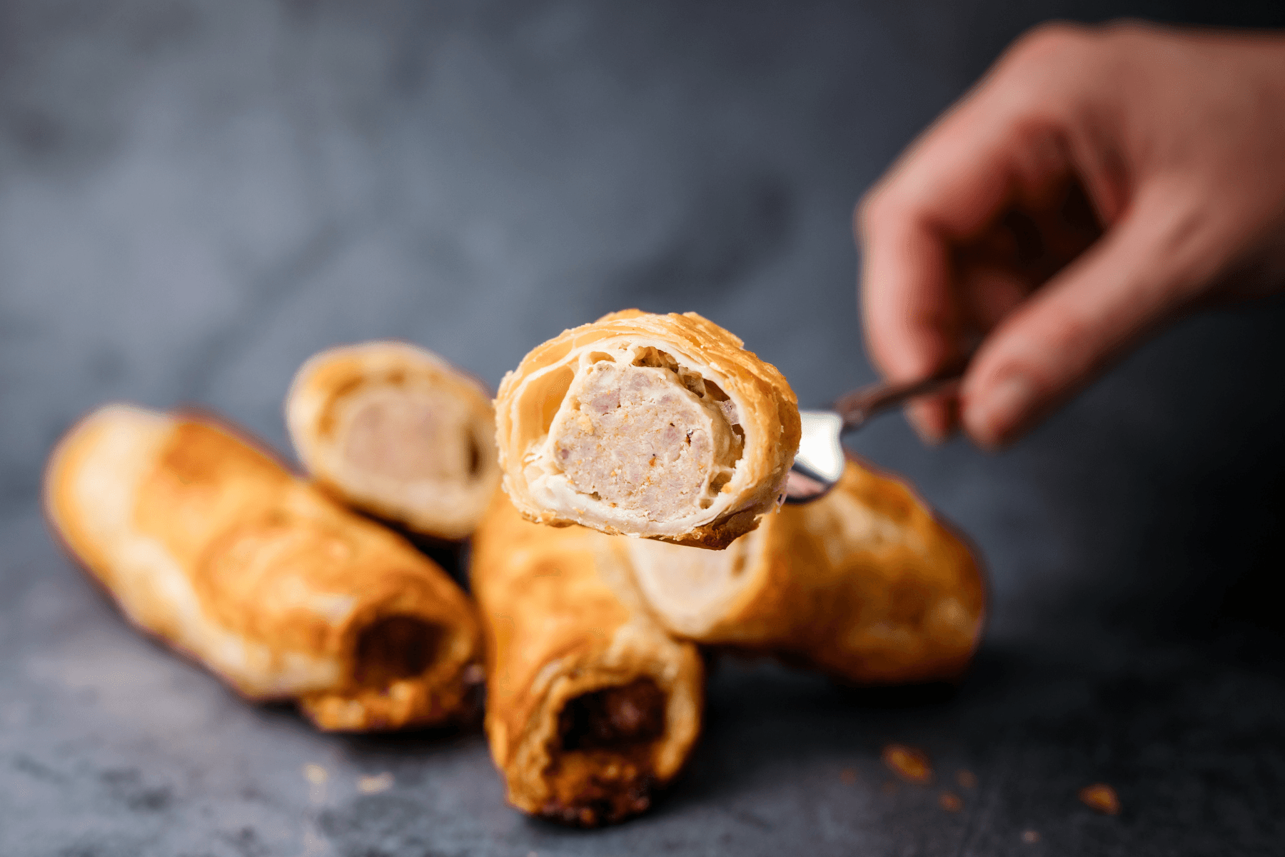 Hand holding a fork with a sausage roll, with two more sausage rolls in the background on a dark surface.