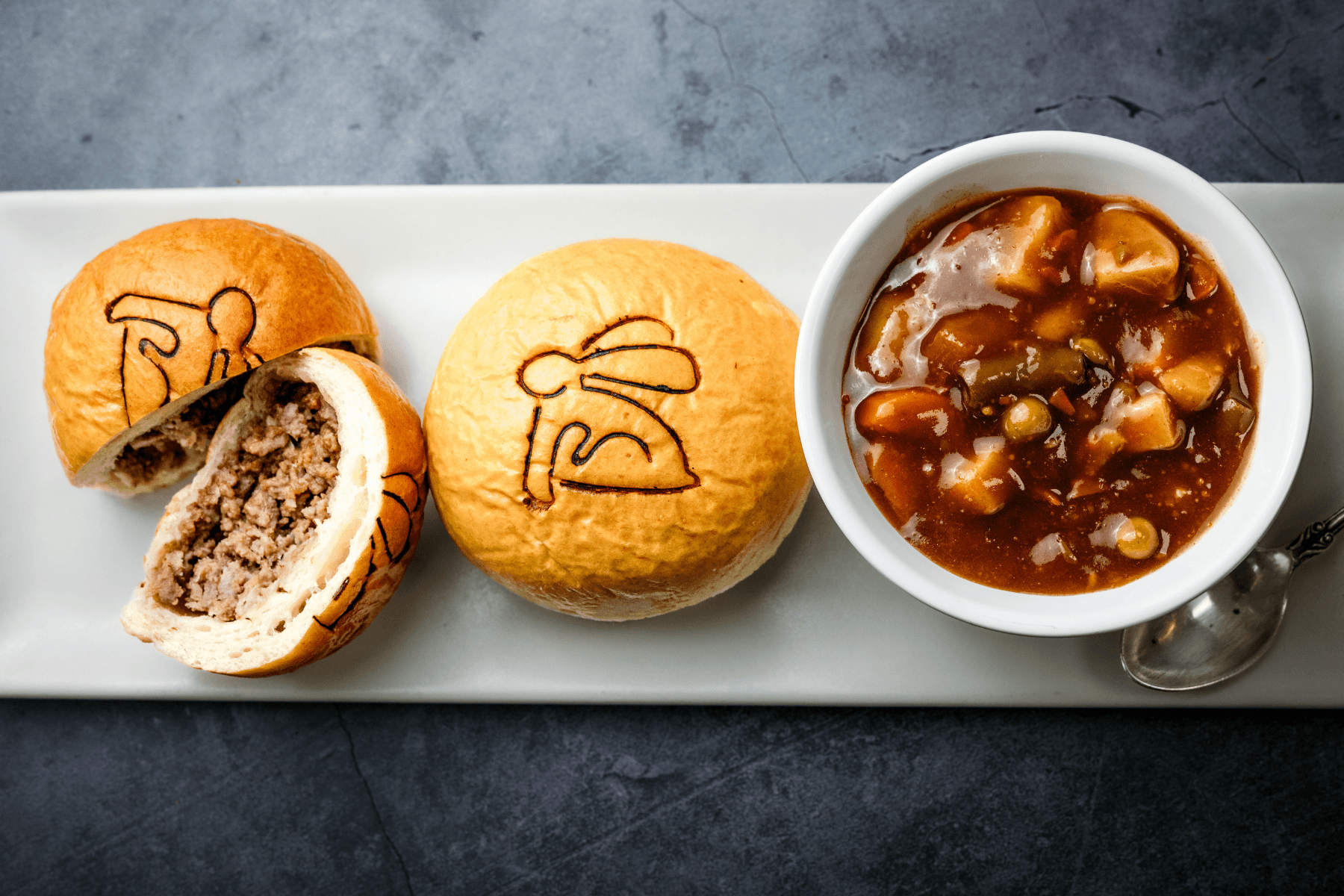Three baked buns with filling and a bowl of stew on a white rectangular plate, on a dark surface.