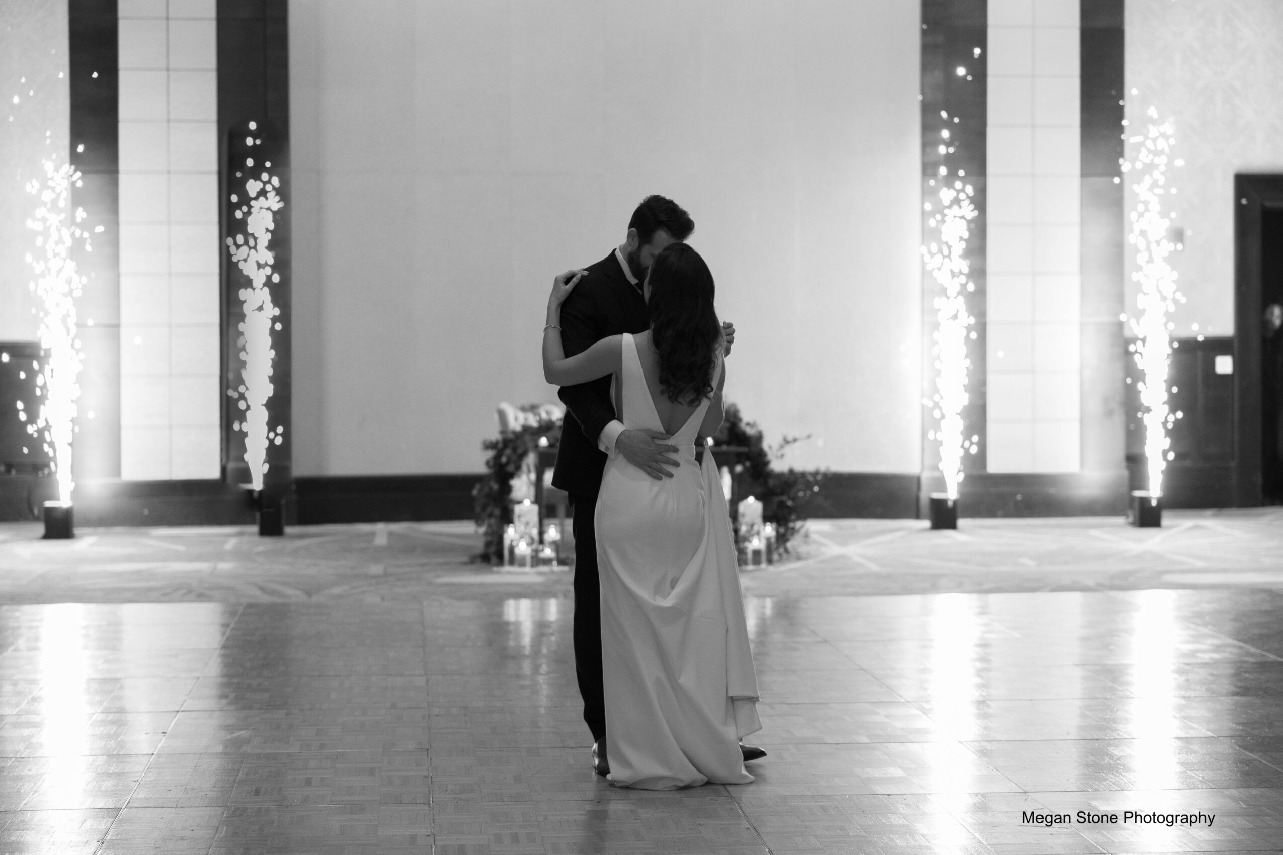 A couple dancing in a large, decorated indoor space with vertical light displays and floral arrangements, black and white photo.