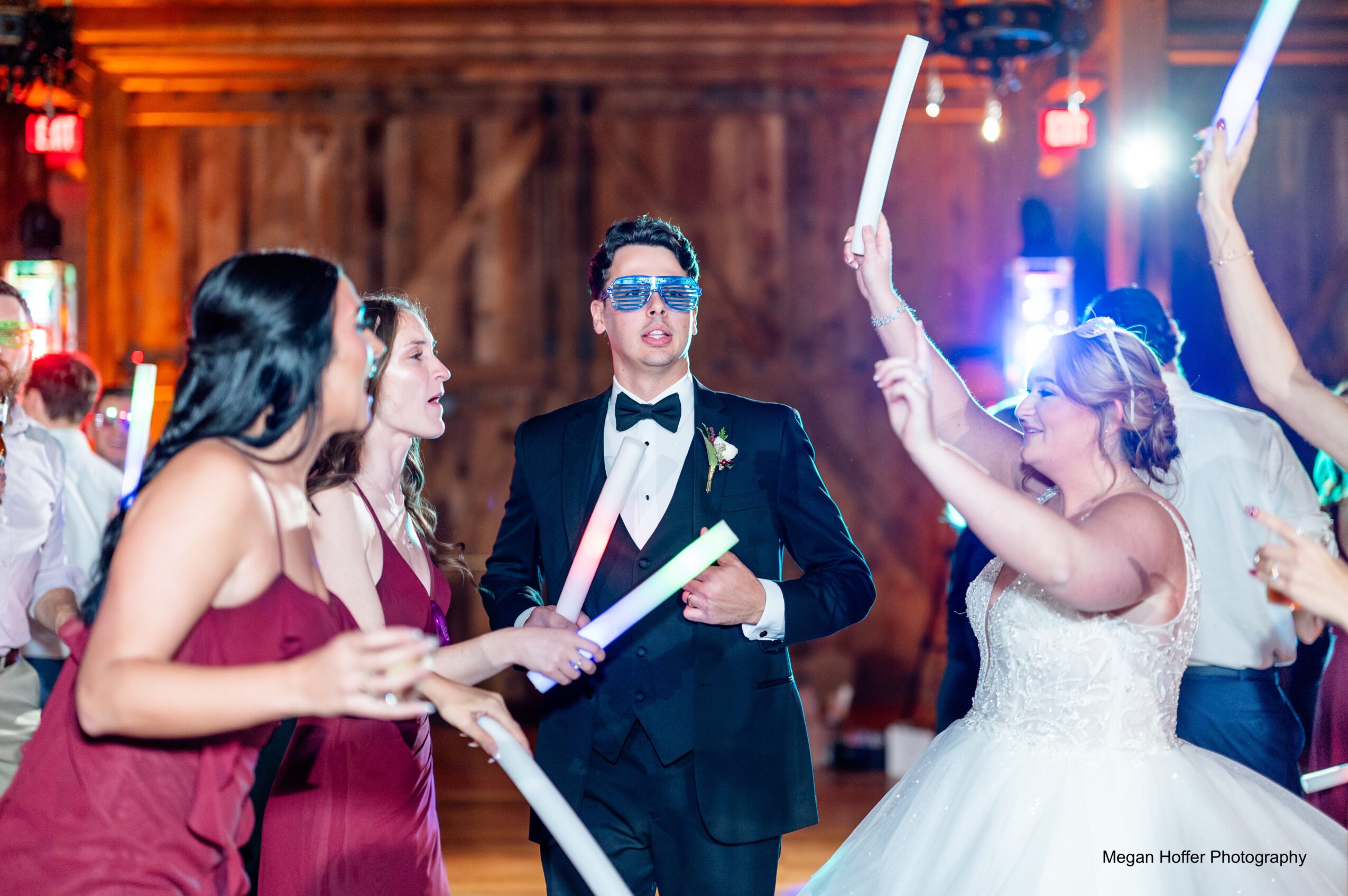 People dancing at a wedding reception, some holding glow sticks, with a wooden background and colorful lighting.