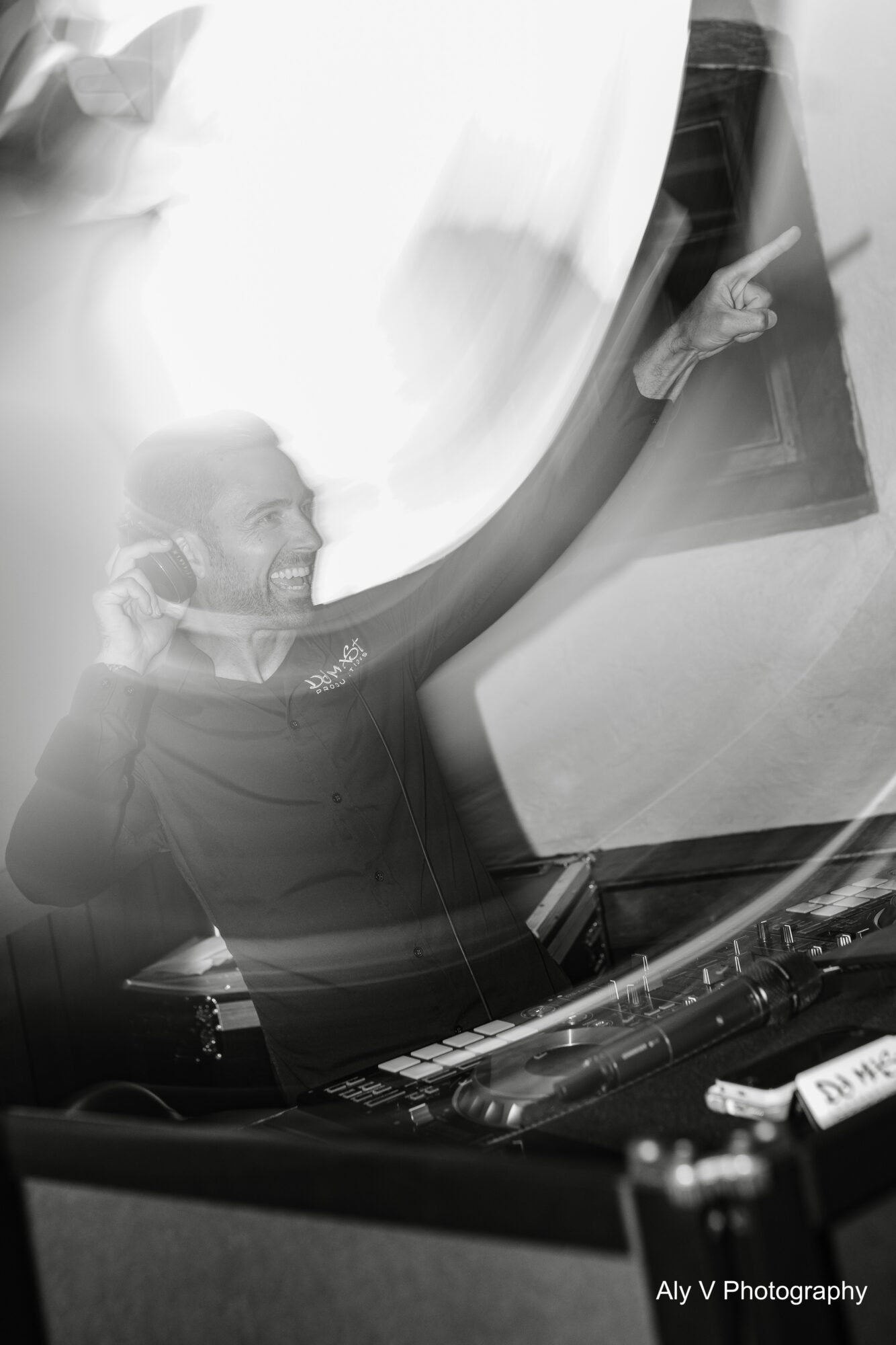 Person smiling, wearing headphones, reflected in a curved mirror or glass, with a DJ or music equipment nearby.