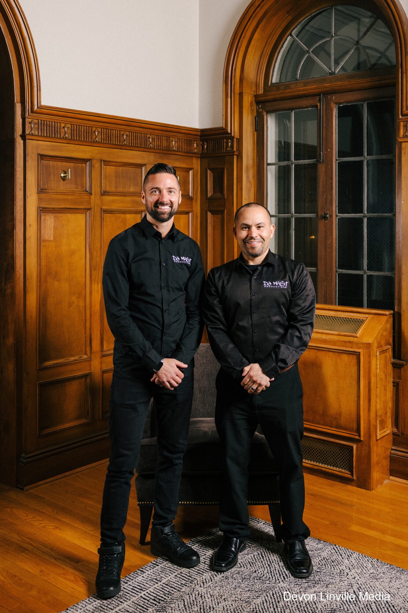 Two men in black jackets stand in a wood-paneled room with large arched window behind them.