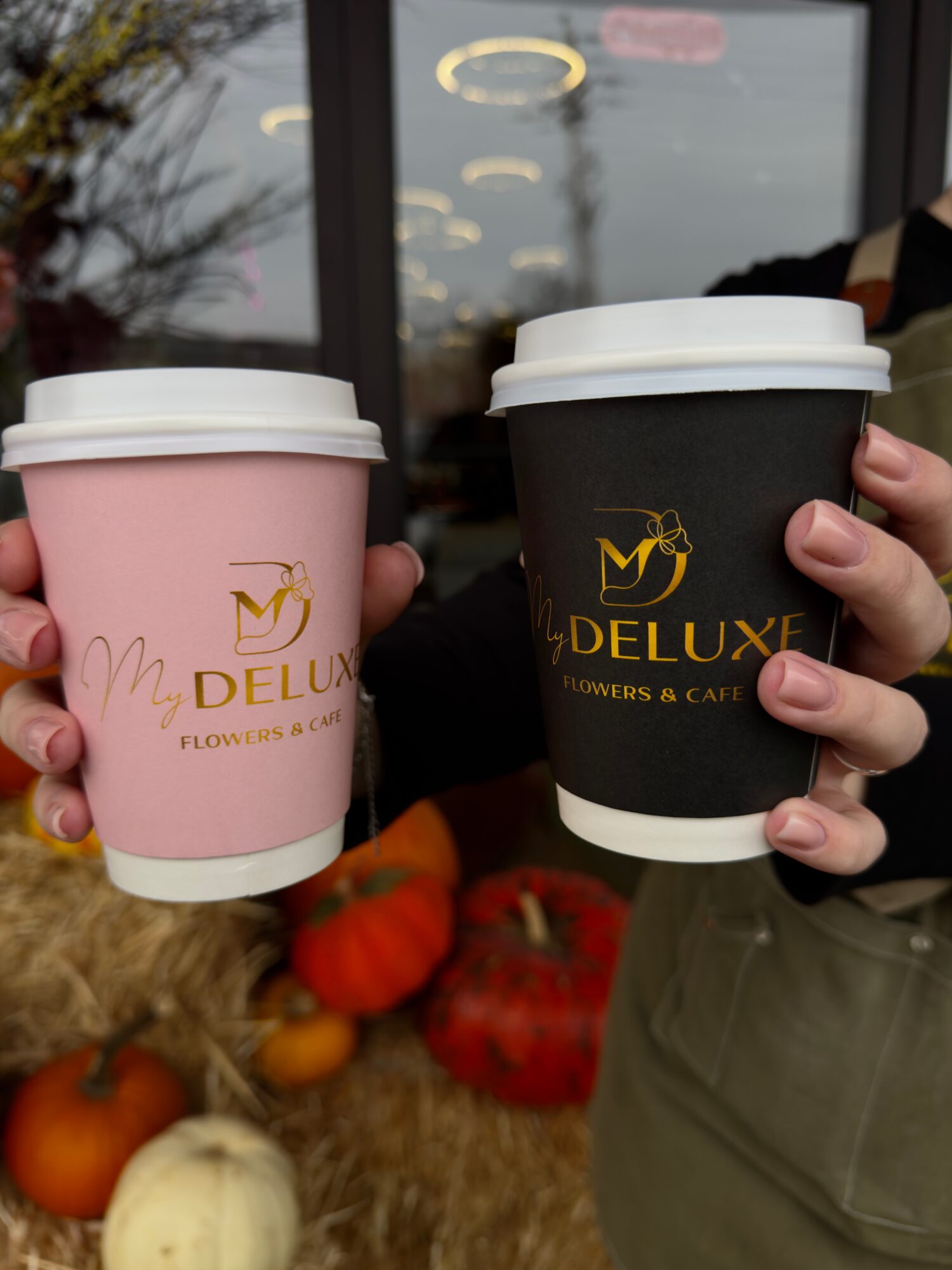 Two people hold coffee cups outside near pumpkins and gourds, with a window reflecting trees and sky in the background.