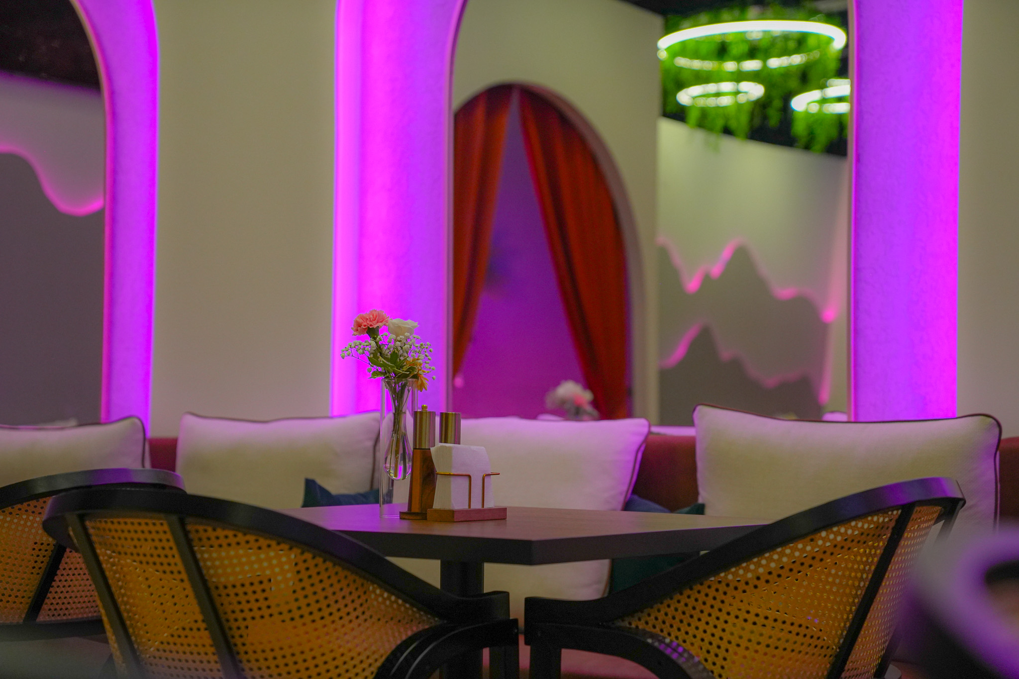 A dining table with chairs in a room with purple and pink lighting, a vase with flowers, and a curtain in the background.