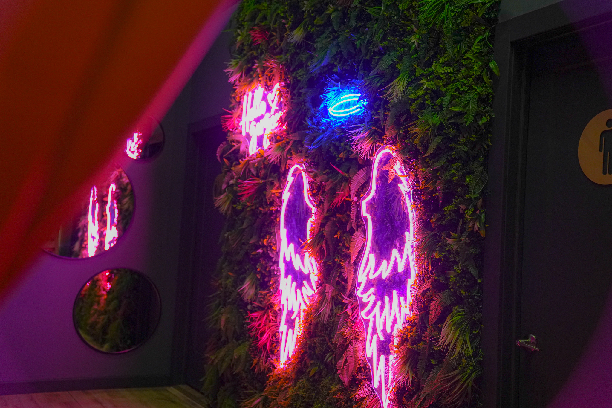 Neon angel wings and halo on a green leafy wall, with a mirror and door nearby.