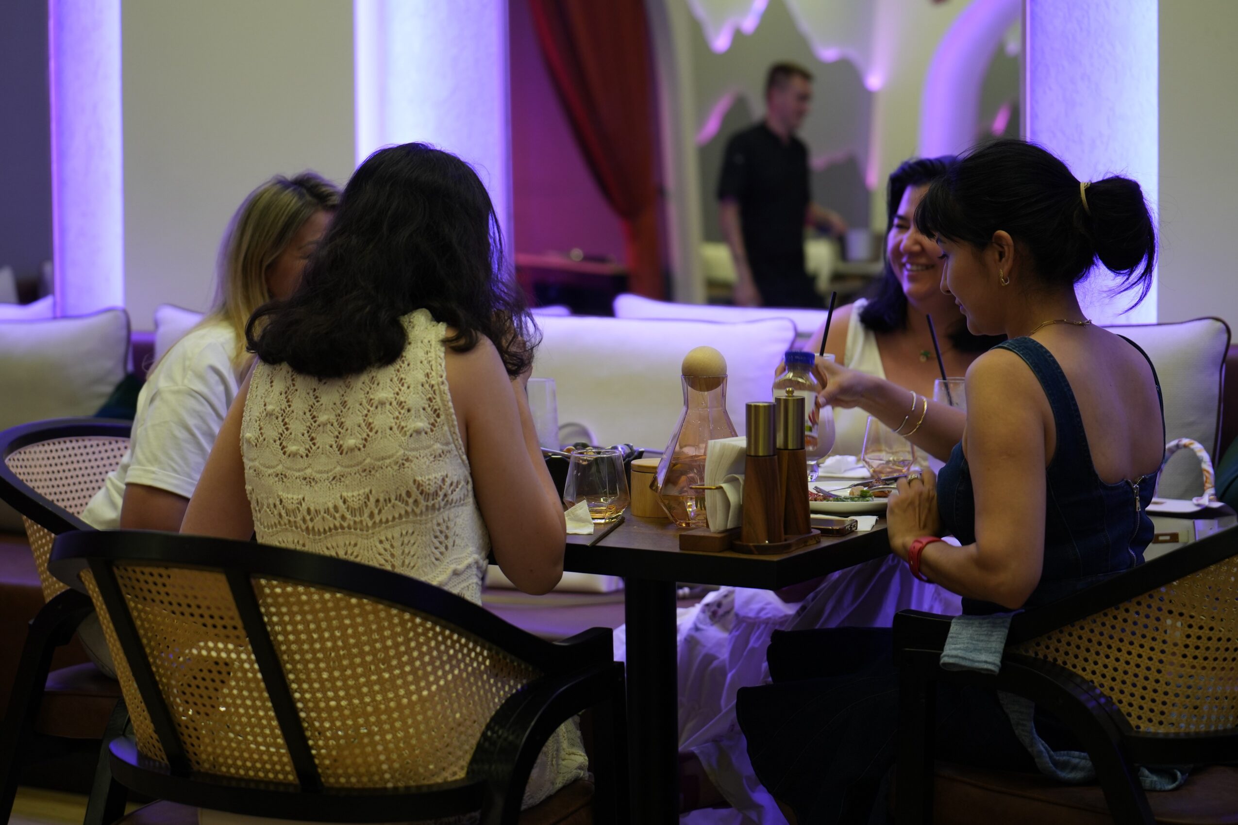 Four women sit around a table in a restaurant, engaging in conversation with drinks and condiments on the table.