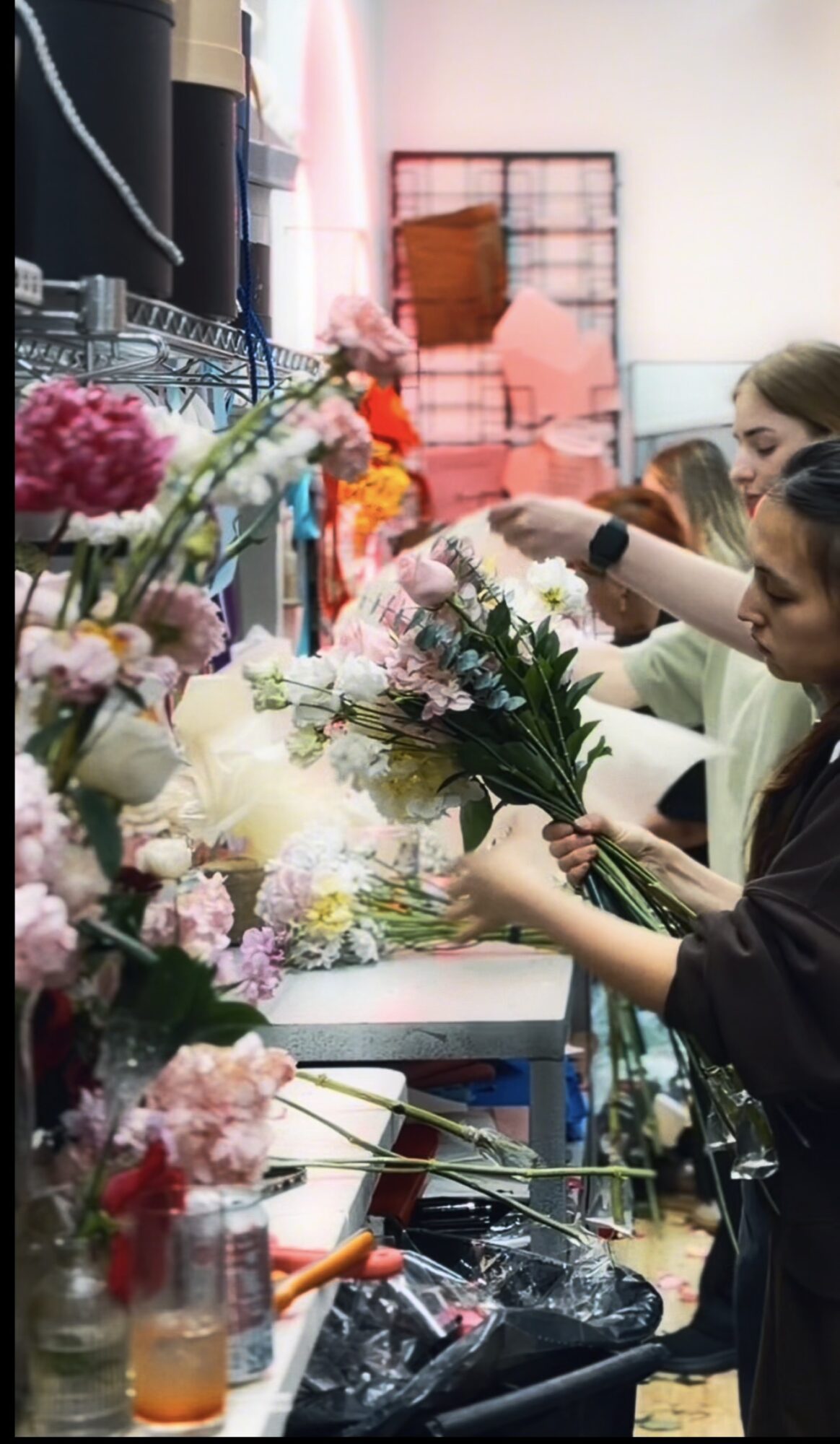 People arranging colorful flowers on a table in a workshop or store, with shelves and boxes in the background.