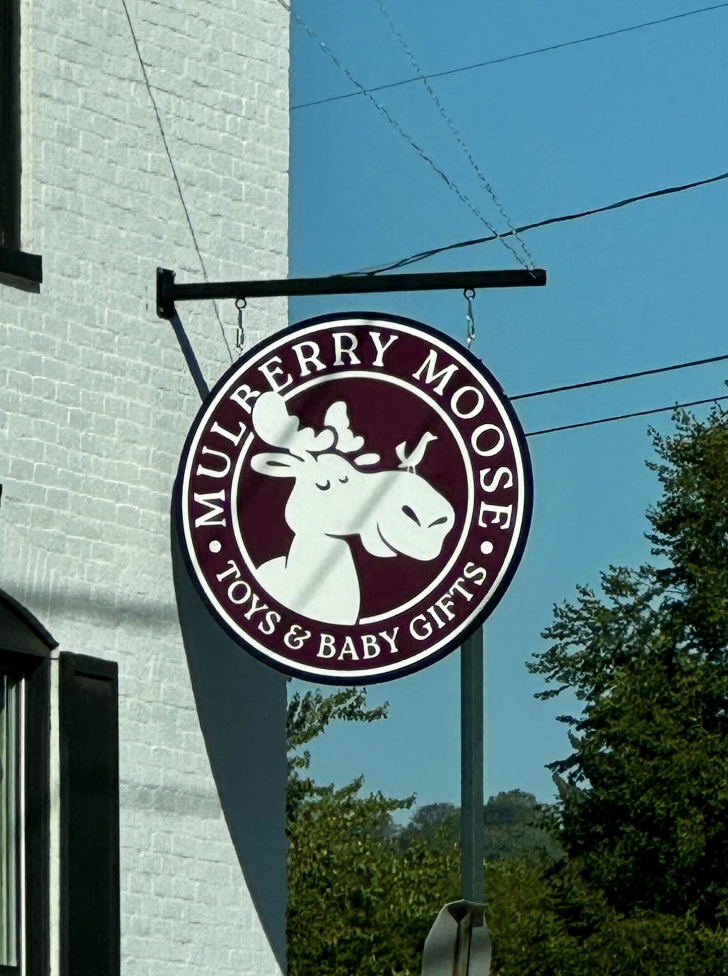 Round sign with a moose illustration and text for Mulberry Moose, Toys & Baby Gifts, hanging outside a building.