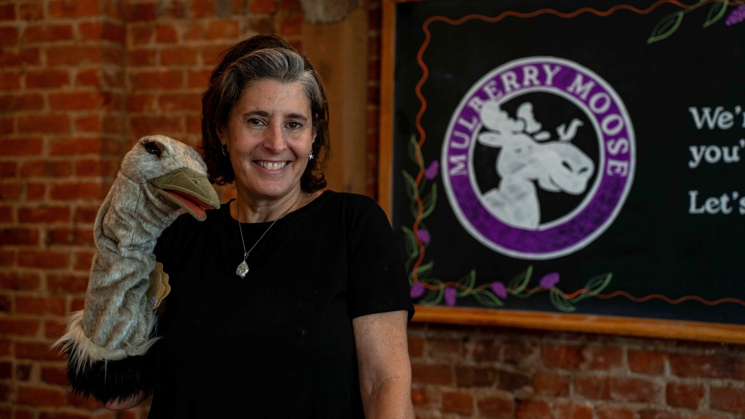 Woman smiling with a puppet hand on her shoulder, standing in front of a brick wall and a sign with a mouse logo.