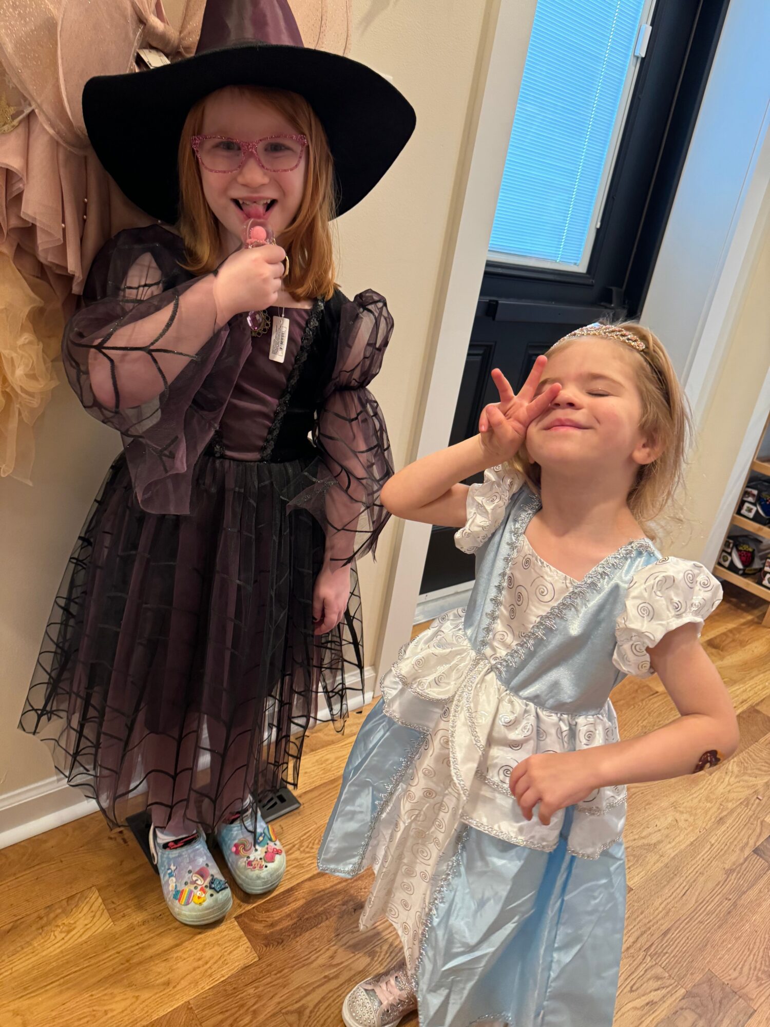 Two children in princess costumes standing indoors, one in a light blue dress and the other in a black dress with a hat, smiling and posing.