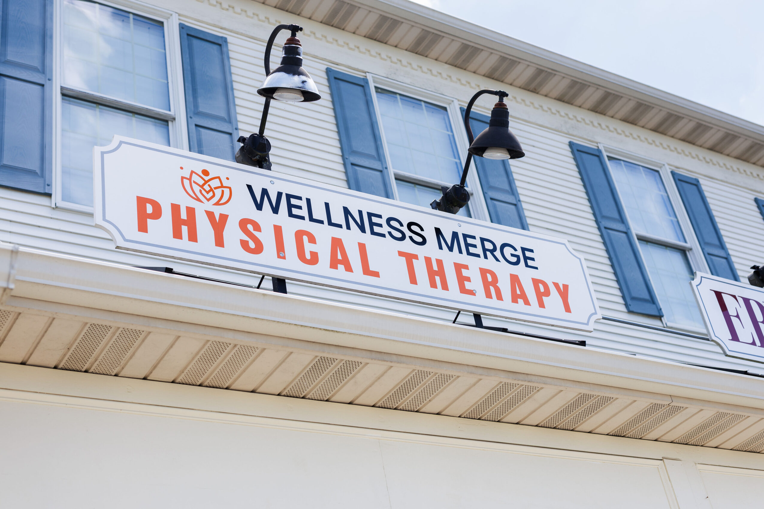 Sign for wellness center offering physical therapy, mounted on a building with blue shutters, under a cloudy sky.