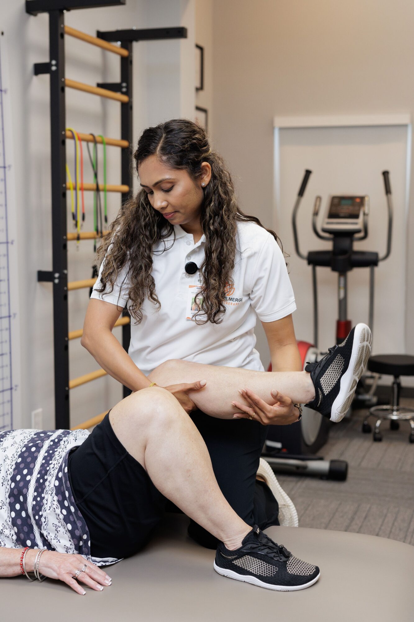 Physical therapist assisting a person with leg exercises in a gym setting, with exercise equipment in the background.