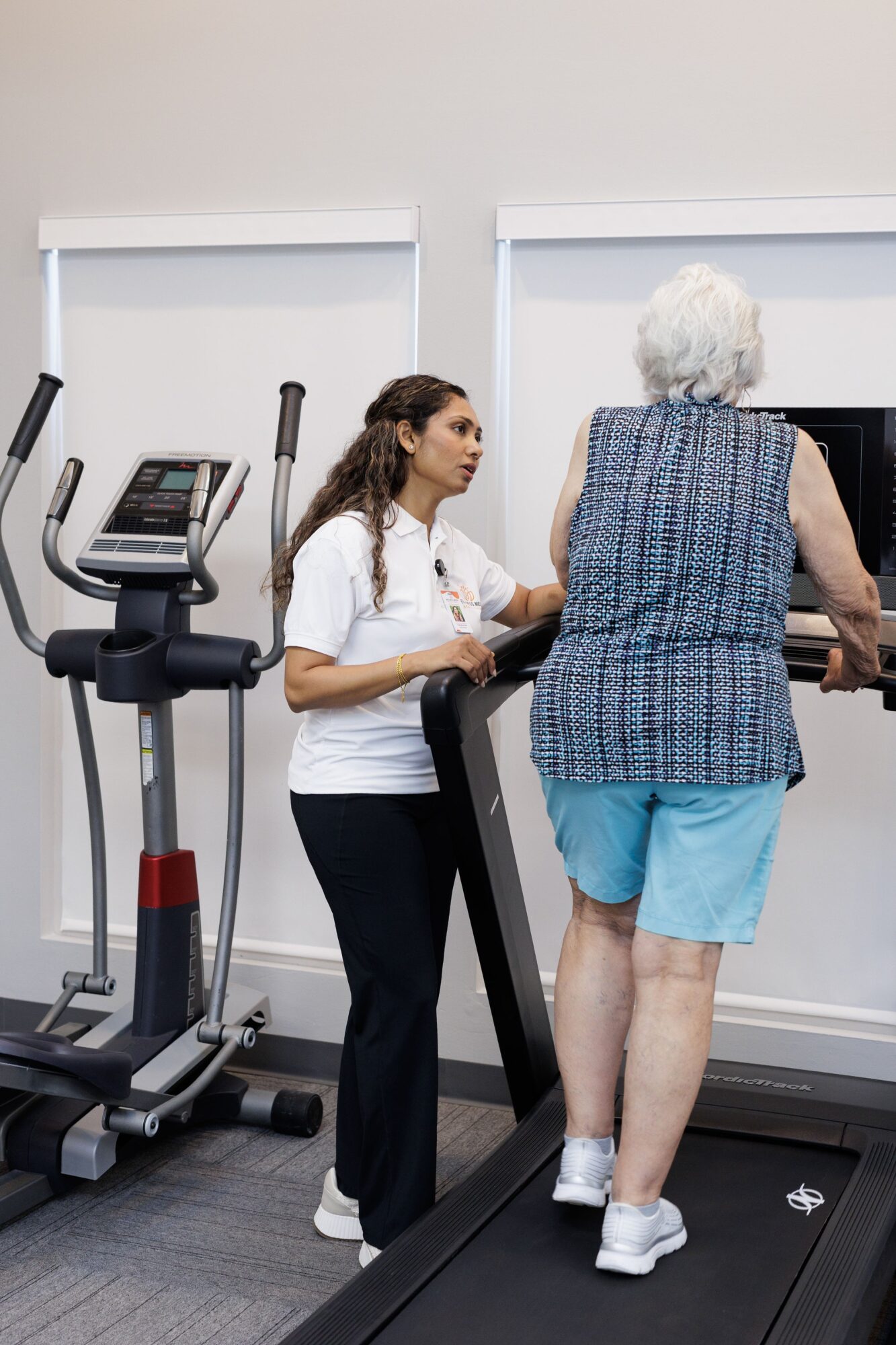 A young woman assisting an elderly woman on a treadmill in a gym, with exercise equipment nearby.
