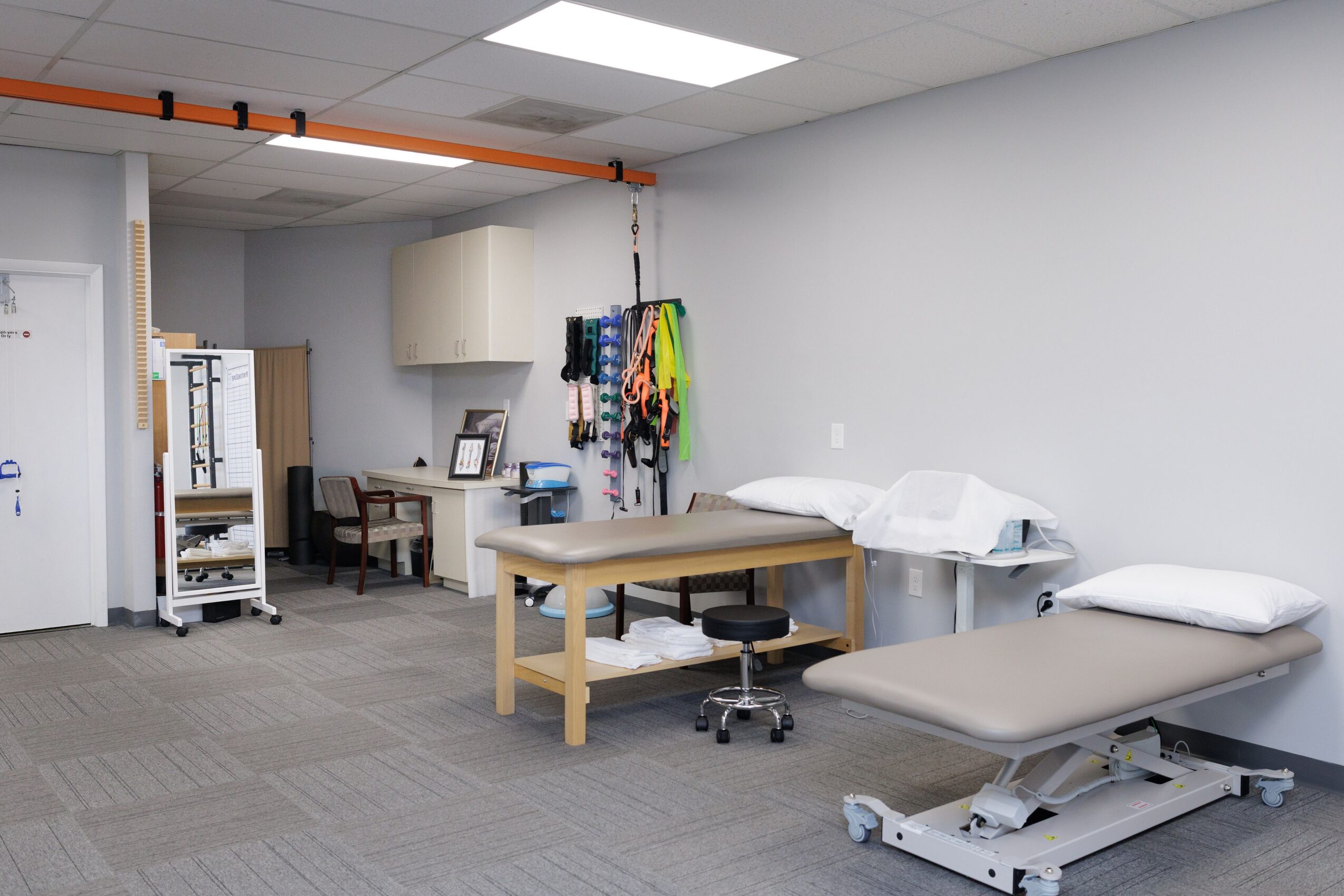 Medical examination room with examination table, chair, and medical equipment, walls painted light blue, ceiling with fluorescent lights.
