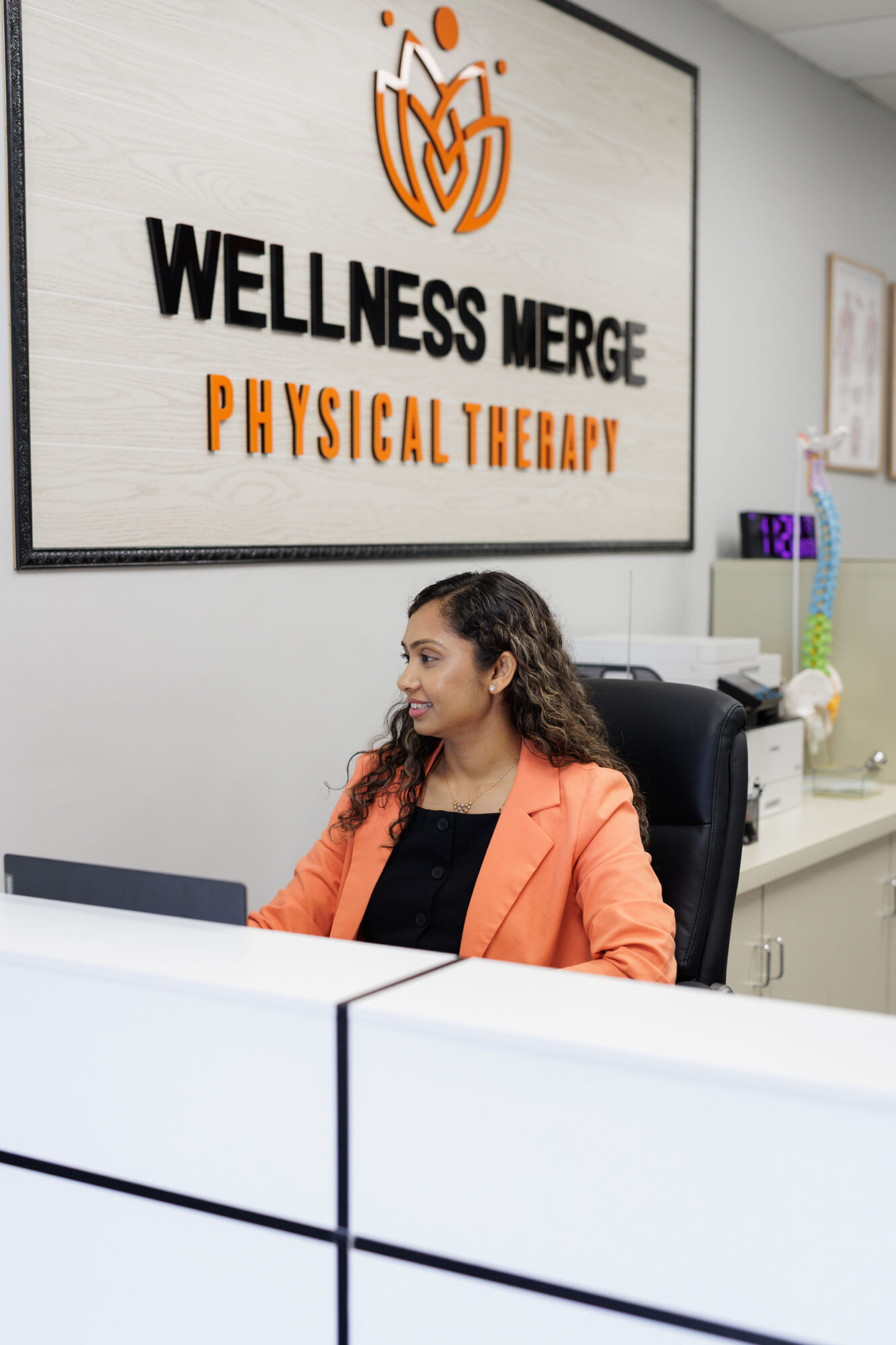 Woman with curly hair in an orange blazer sitting at a desk in wellness center, sign on wall behind her.