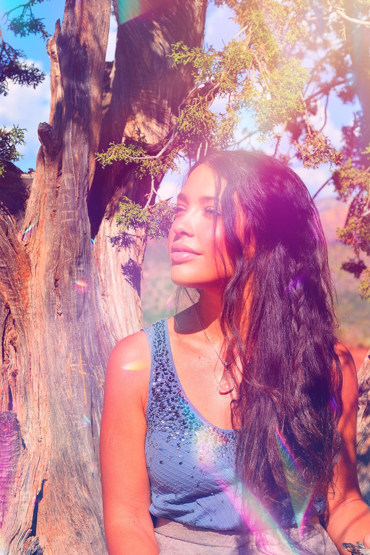 Woman with long dark hair standing outdoors near a large tree, sunlight filtering through leaves, looking to the side.