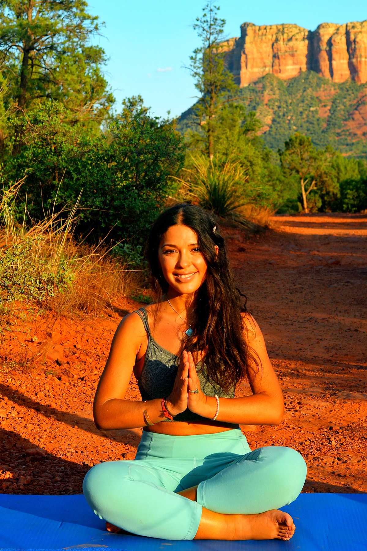 Woman practicing yoga outdoors with a mountain and trees in the background, sitting cross-legged with hands in prayer position, smiling.